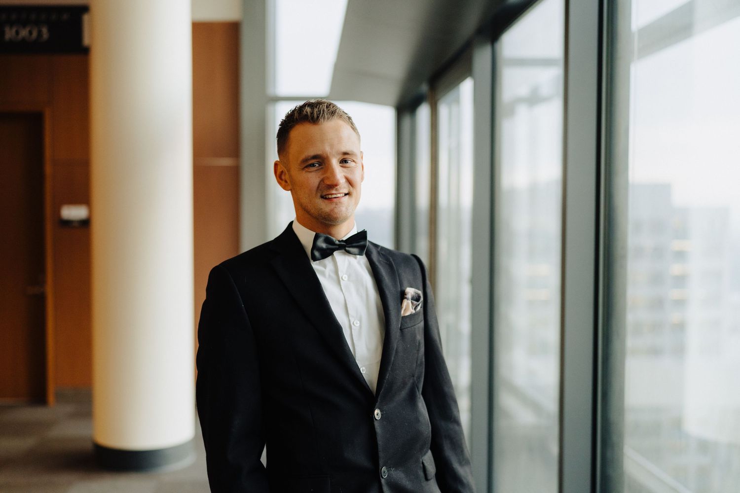 Groom in classic black tuxedo and bowtie poses by window in modern building.