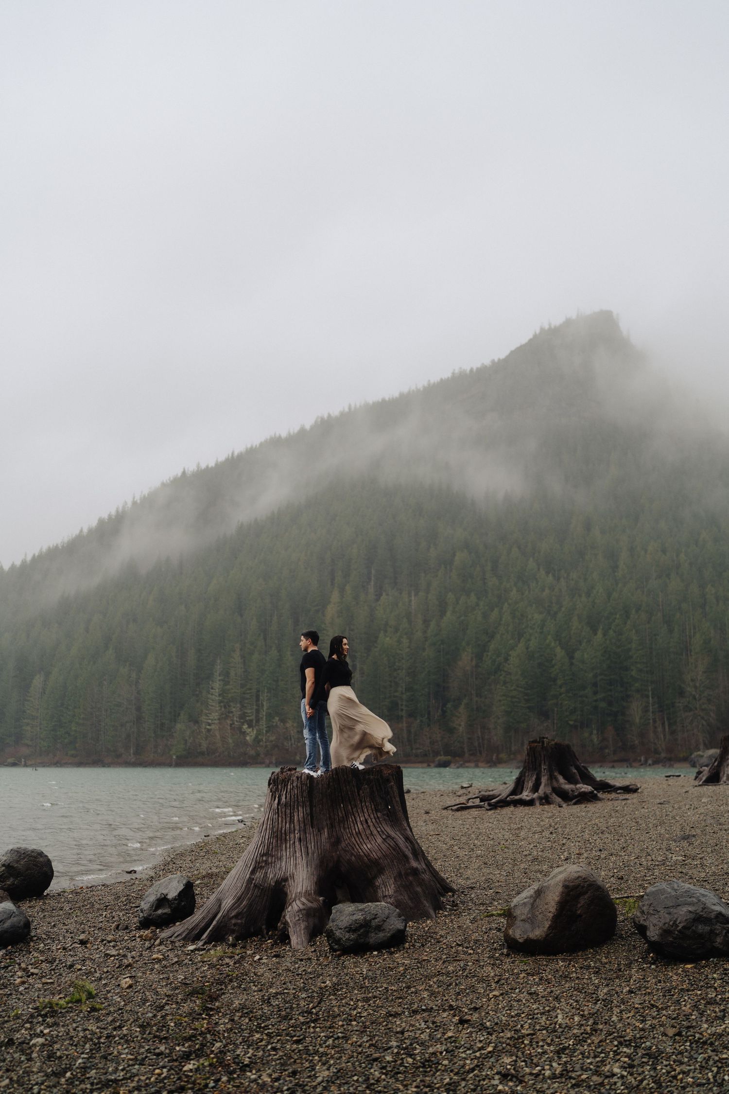 Two people pose on a massive tree stump overlooking a serene mountain lake surrounded by fog.
