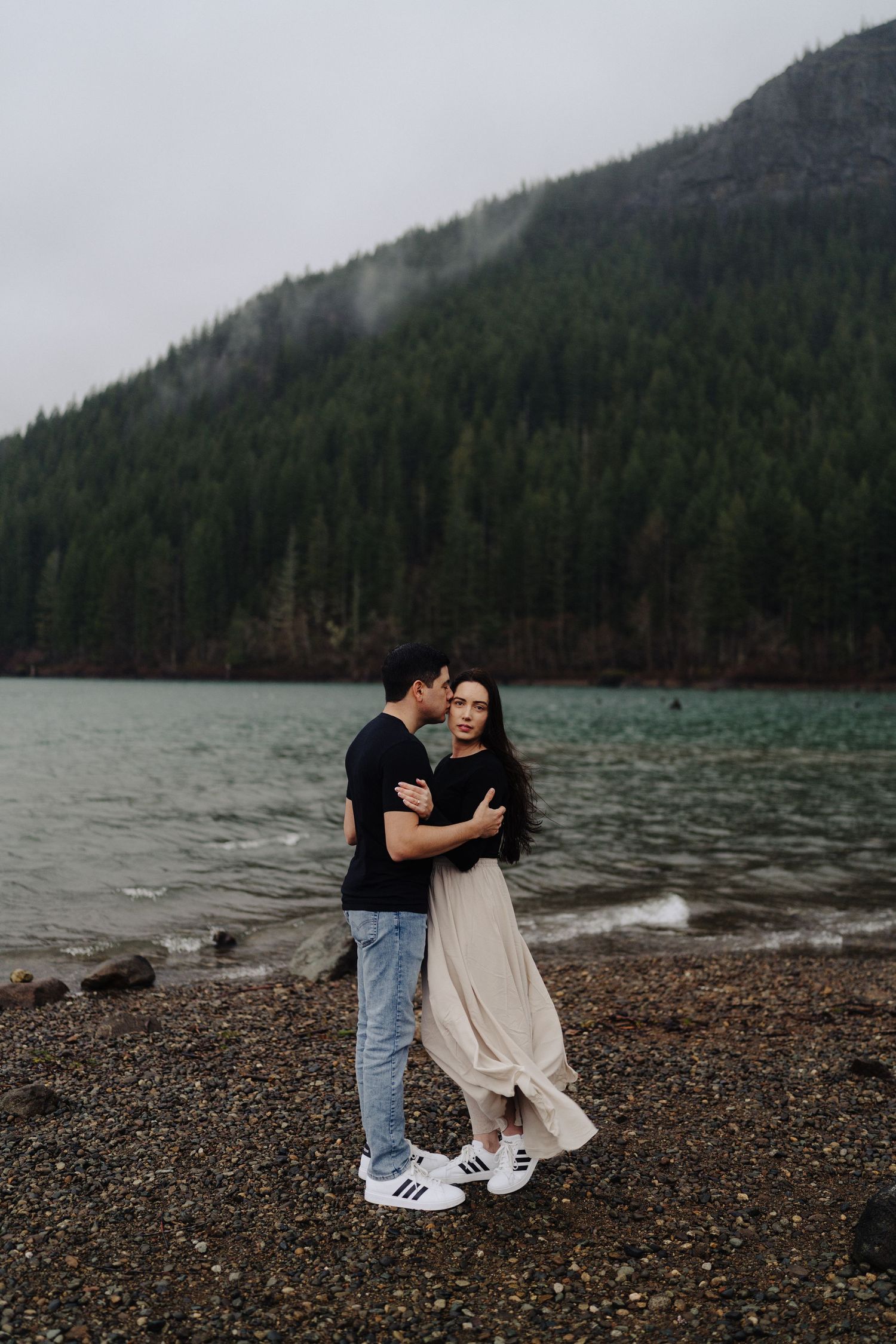 A romantic embrace captured on a rocky shoreline with misty mountain peaks in the background.