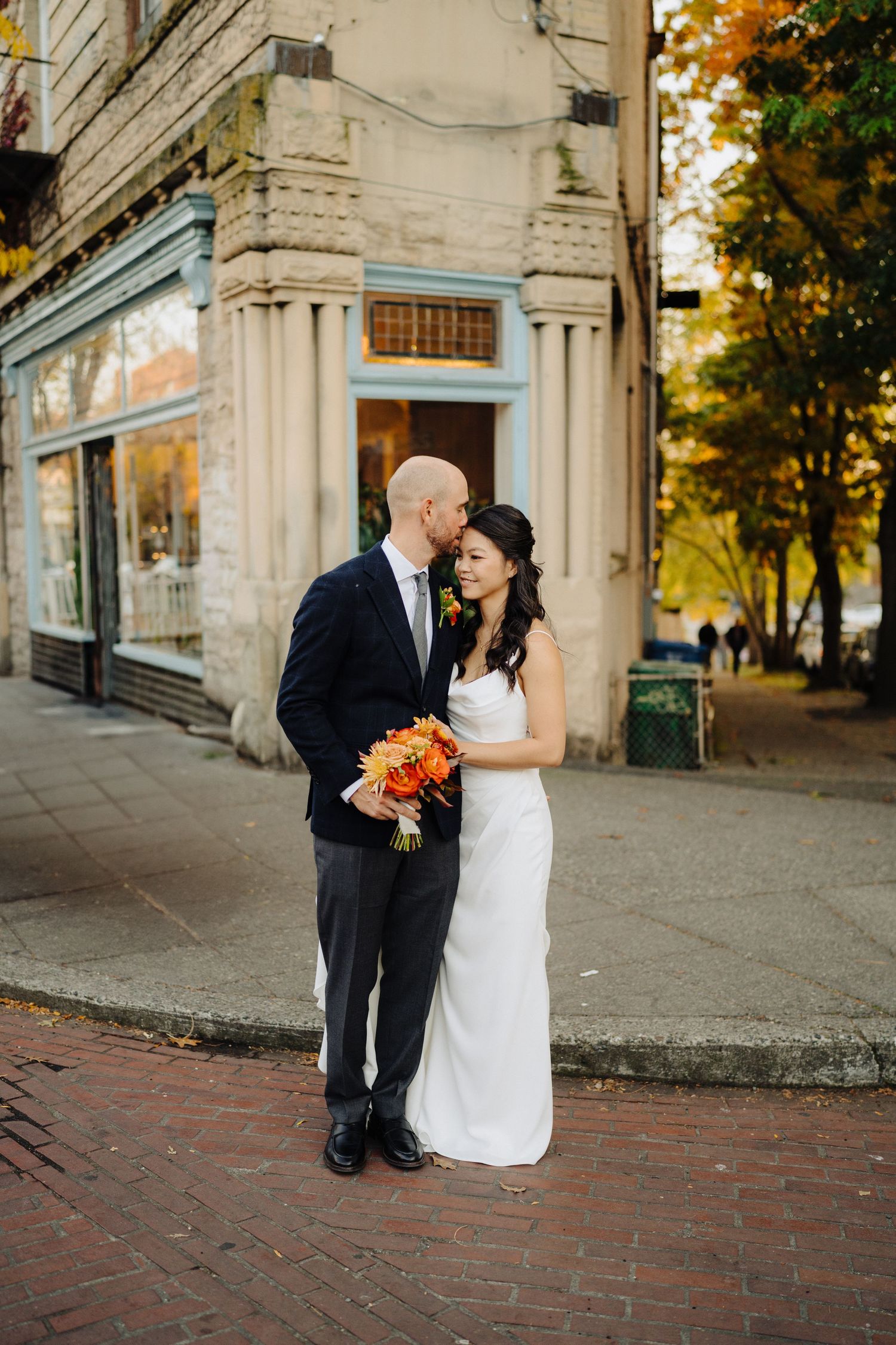 Wedding couple posing outside historic building with orange bouquet on brick sidewalk.