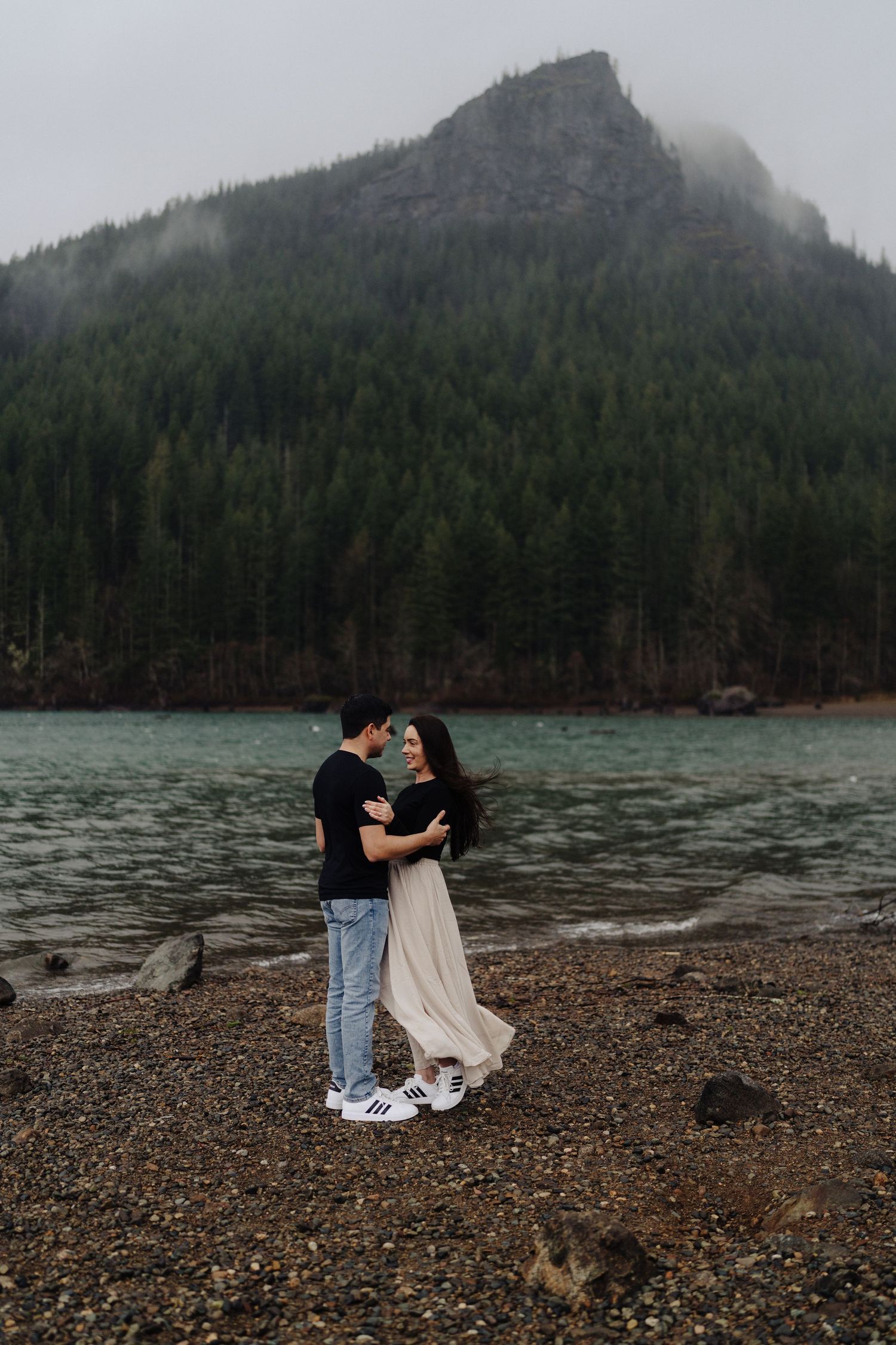A couple shares an intimate moment on a rocky shoreline with a dramatic mountain peak shrouded in fog behind them.