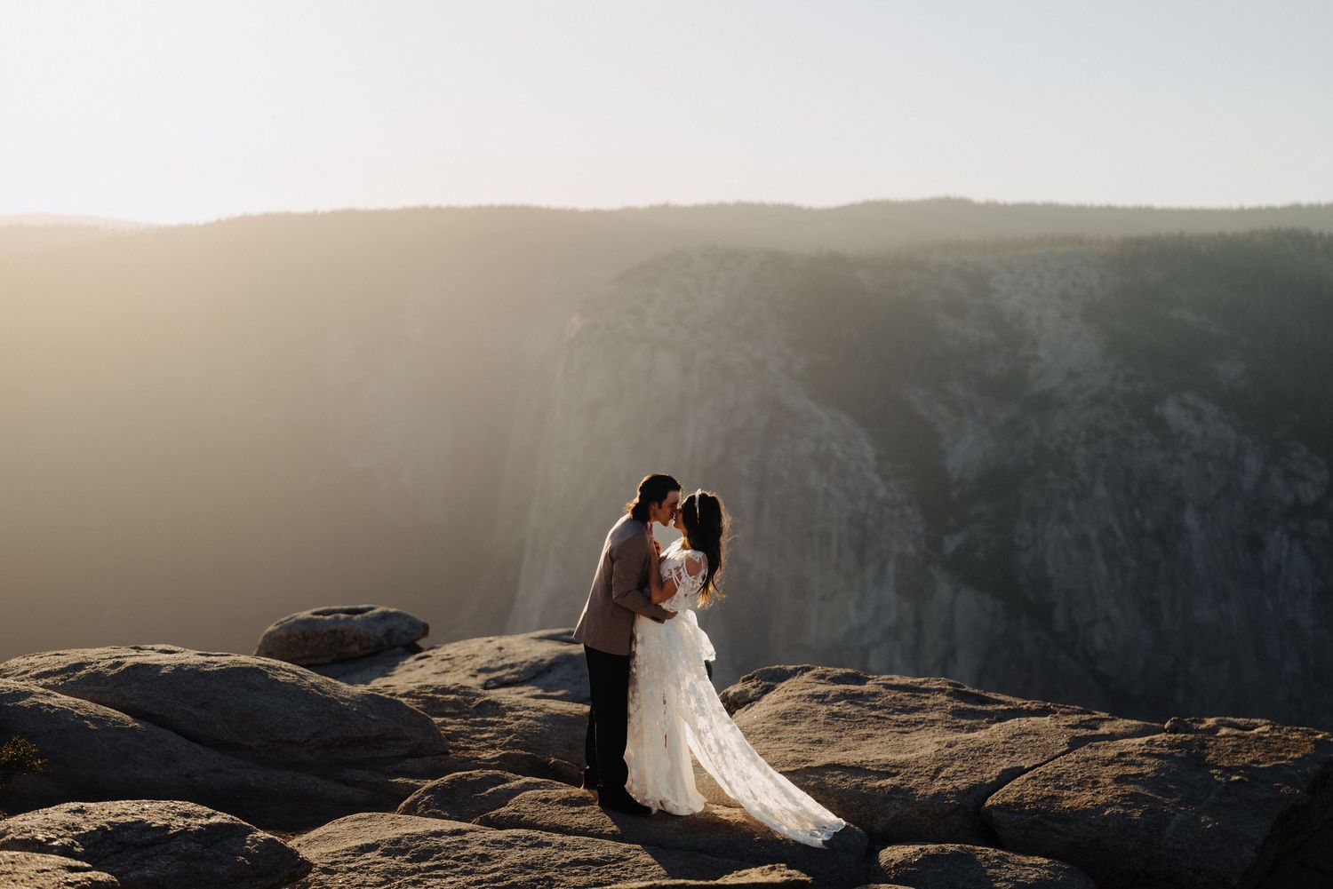 A couple in wedding attire embraces on a dramatic clifftop overlook during golden hour with misty mountains in background.