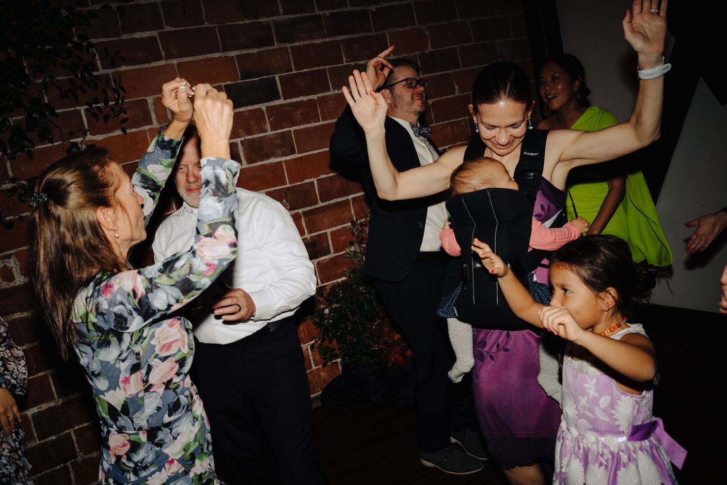 Wedding reception guests dancing and celebrating against brick wall backdrop.