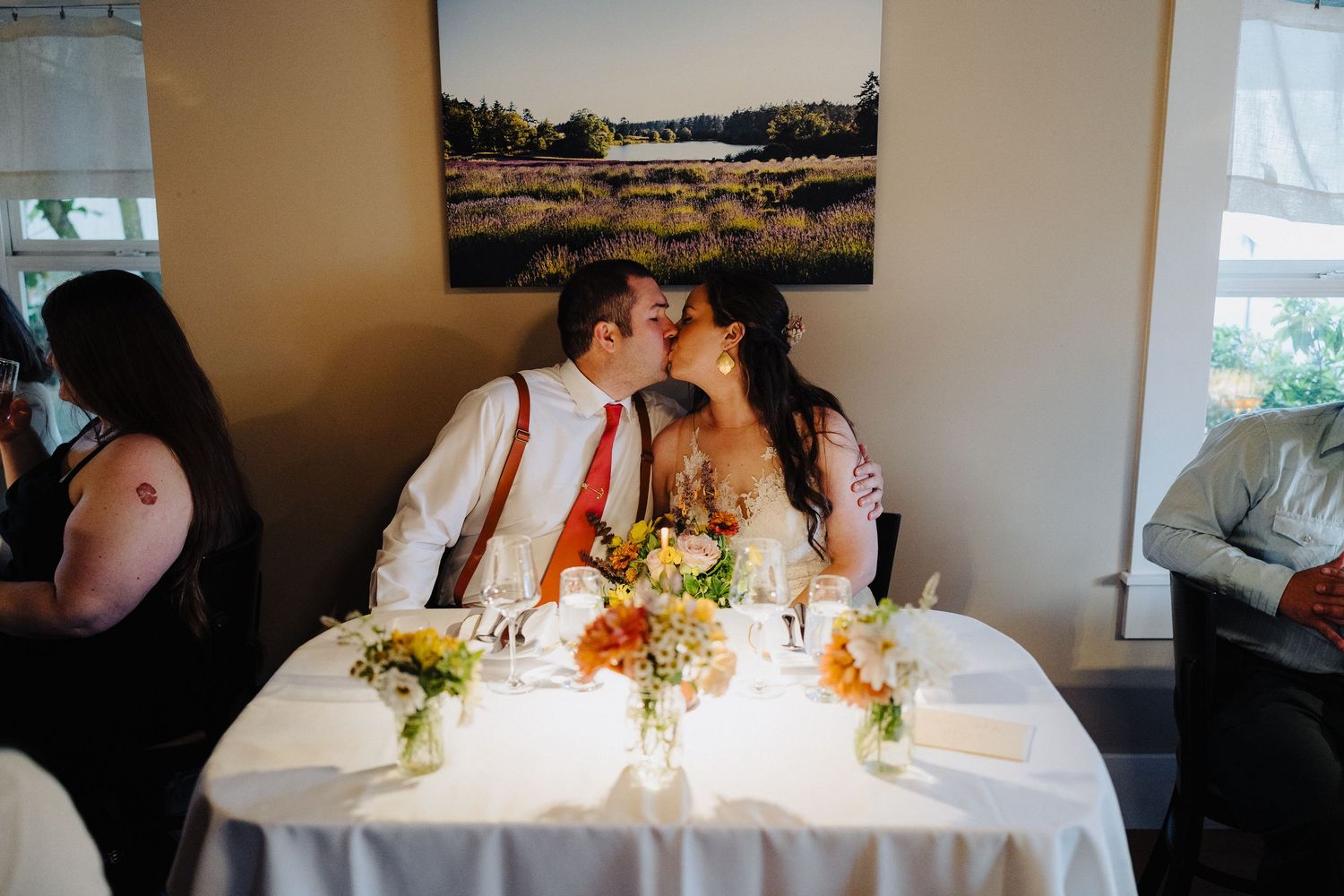 A couple shares a romantic kiss at their wedding reception table decorated with colorful floral centerpieces.