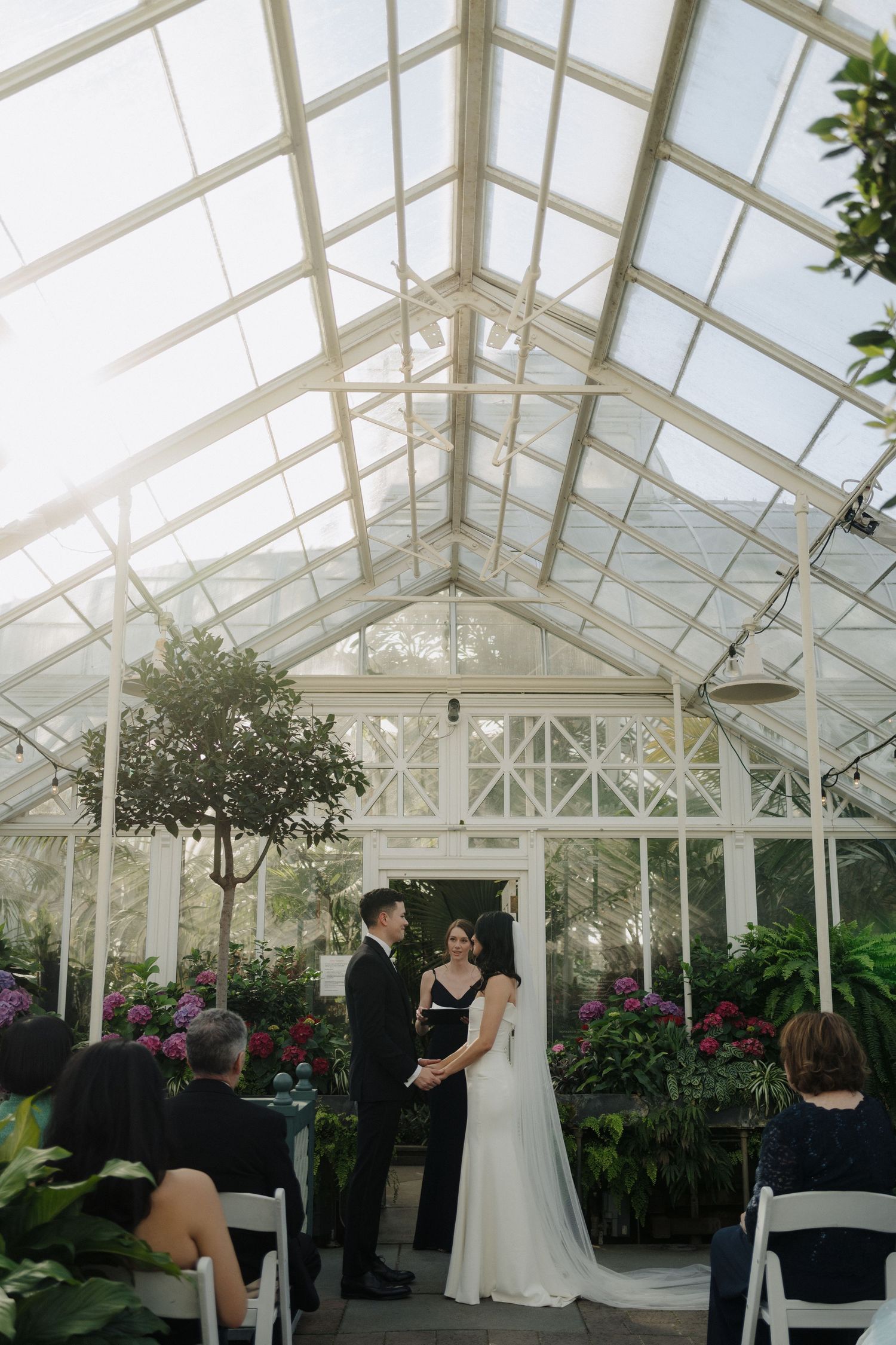 An elegant wedding ceremony unfolds inside a bright greenhouse filled with lush greenery and white folding chairs.