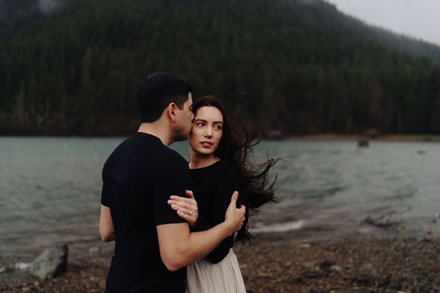 Couple in loving embrace on rocky beach with dramatic mountain and lake landscape.