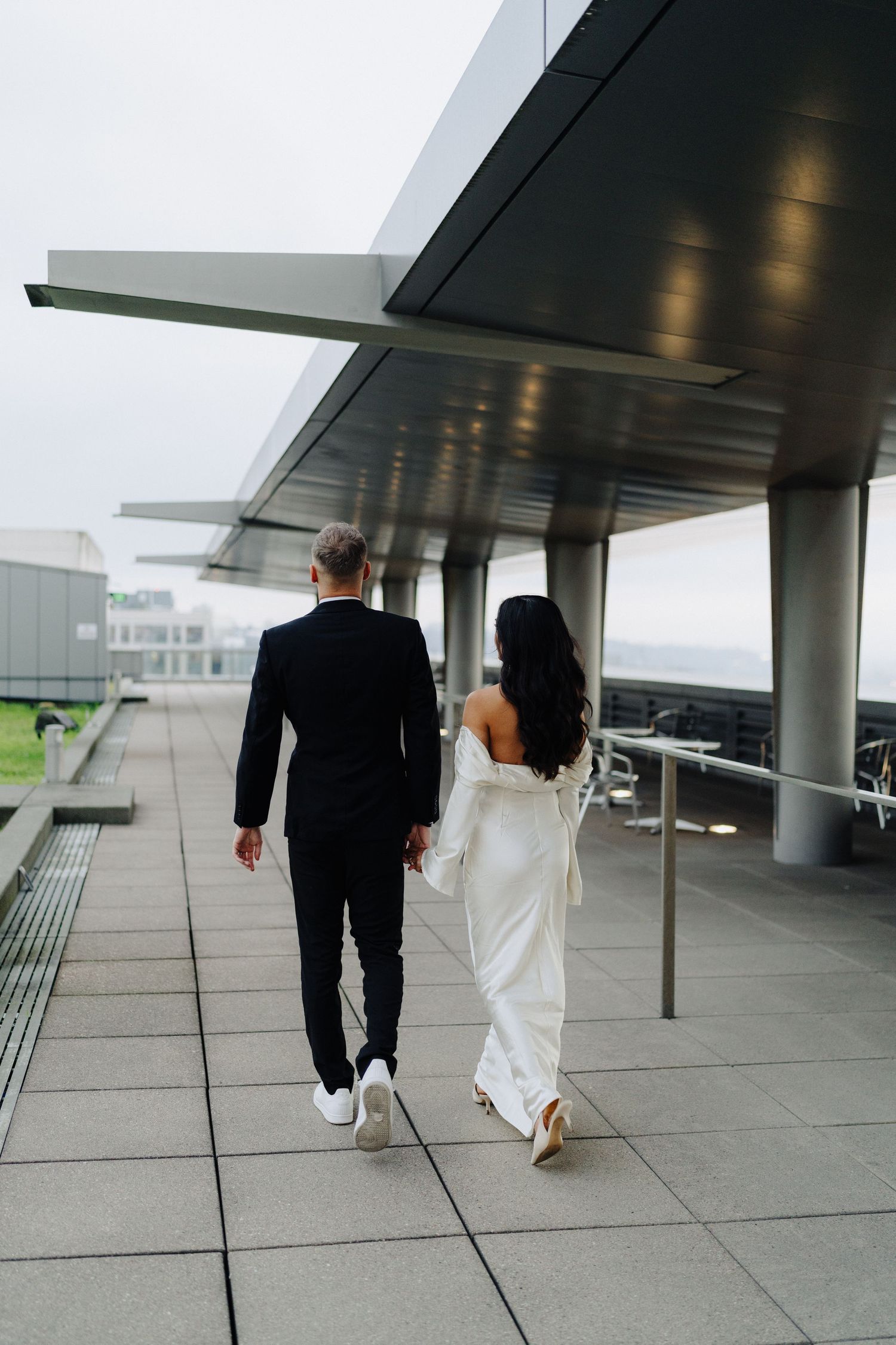 A couple in wedding attire walks along a modern building terrace with architectural details.