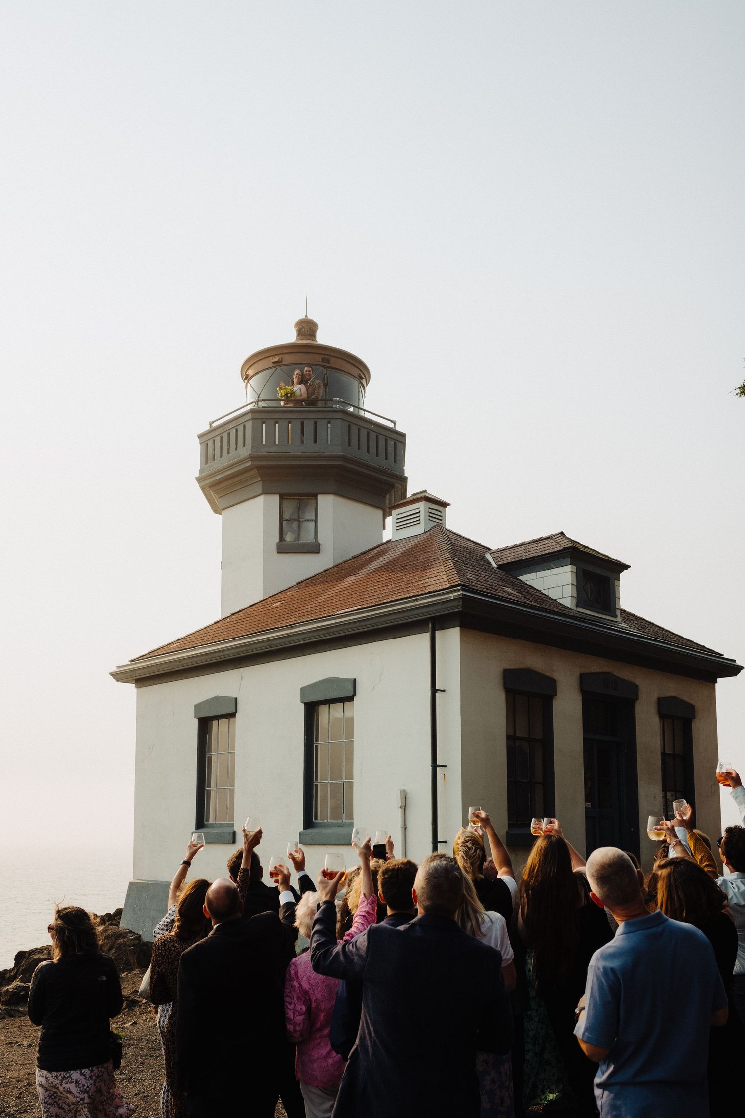 A crowd gathers in front of a white lighthouse during a misty wedding celebration.