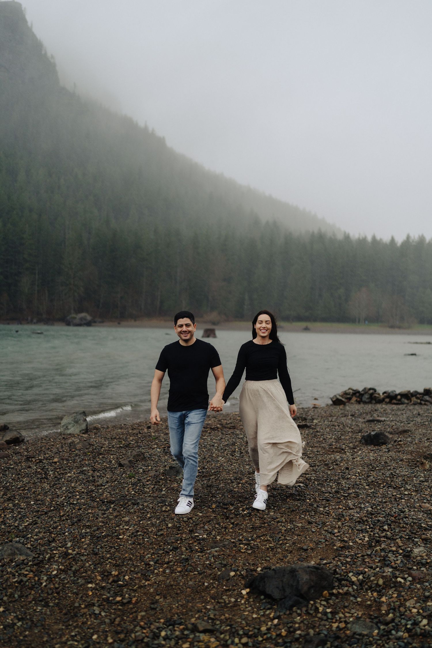 A couple walks hand in hand along a rocky shoreline with fog-covered mountains in the distance.