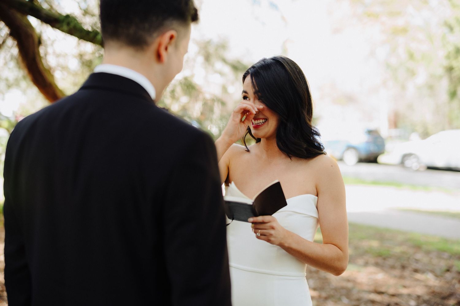 A bride in a strapless white wedding dress reacts emotionally while reading a note outdoors during a wedding ceremony.