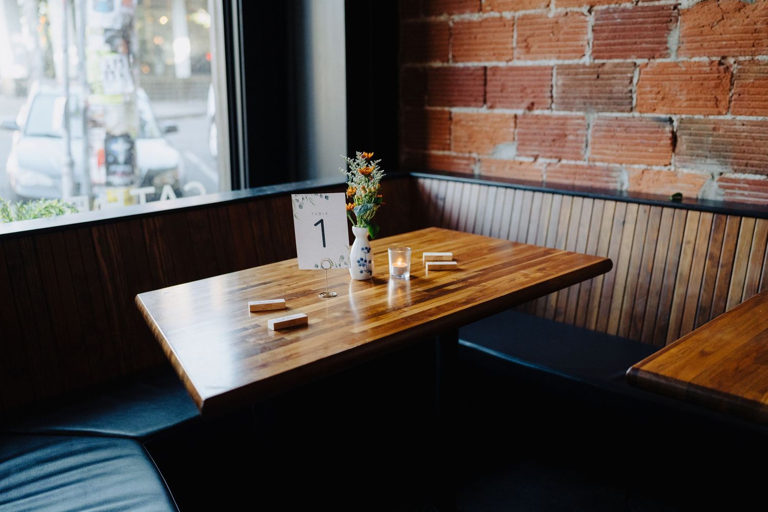 Empty restaurant booth with wooden table and small vase of flowers against brick wall and window.