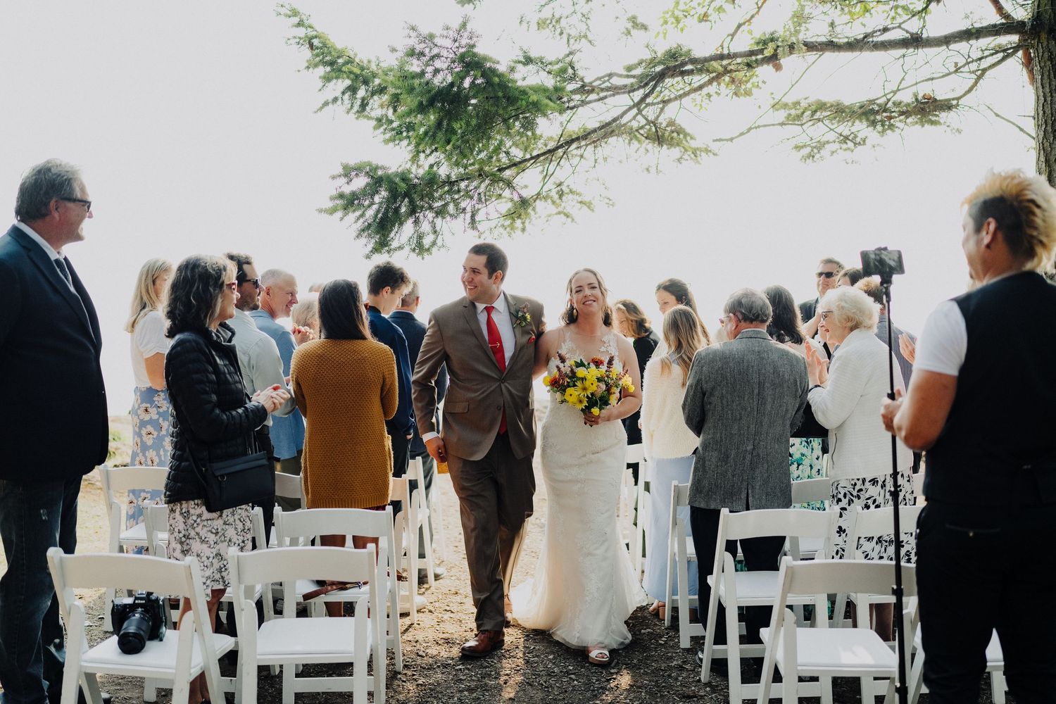 Guests gather outdoors for a wedding ceremony on white chairs with trees and greenery in the background.
