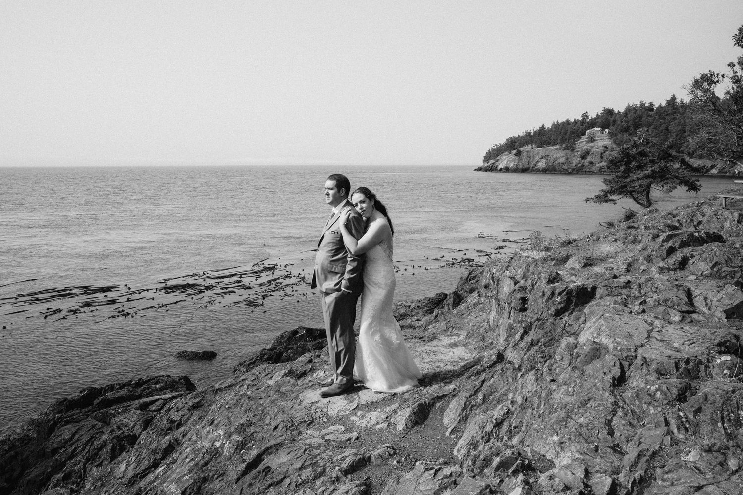 A couple embraces on rocky coastal cliffs with the ocean and shoreline in the background.