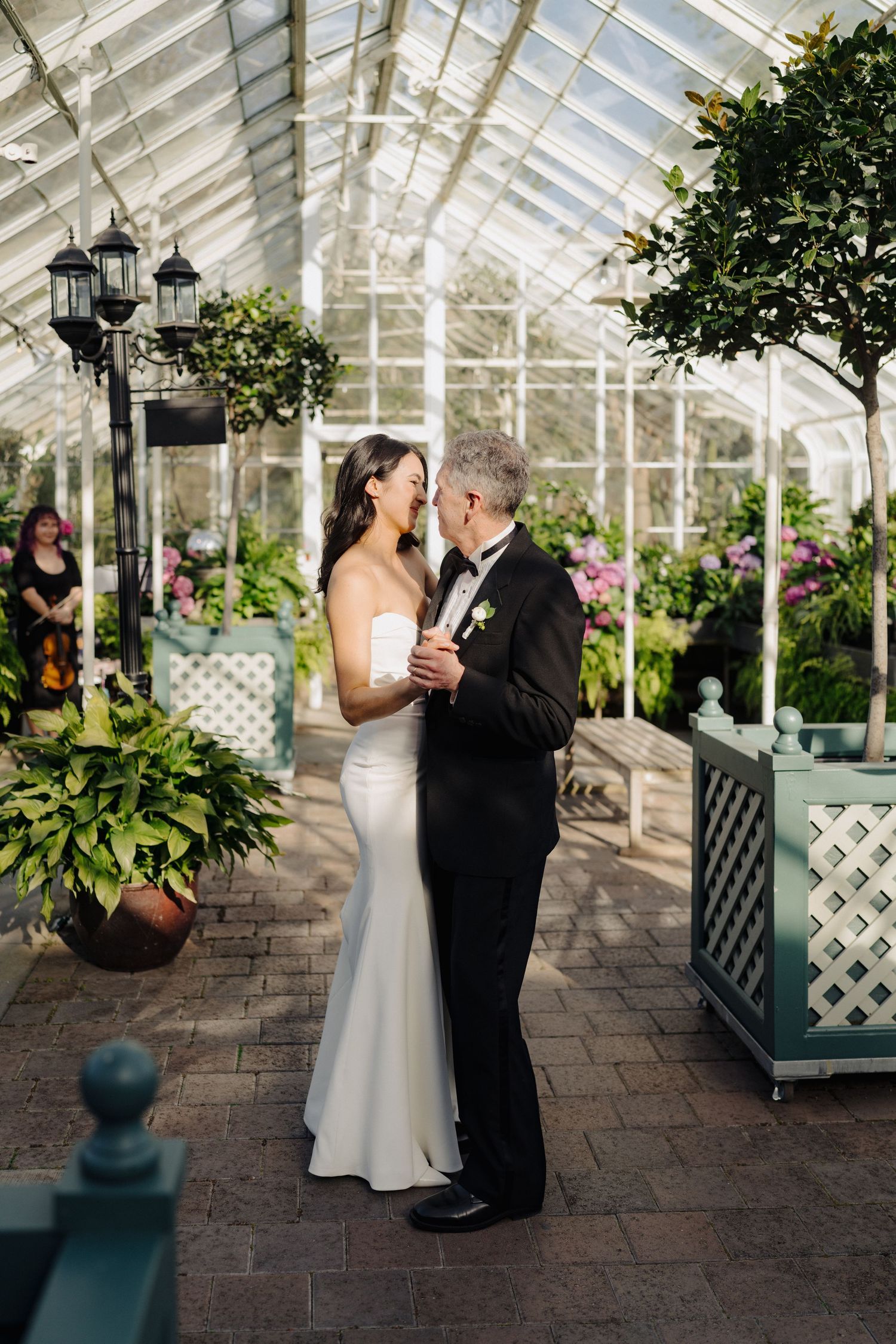 A romantic wedding moment captured in an elegant greenhouse setting with twinkling lights and tropical plants.