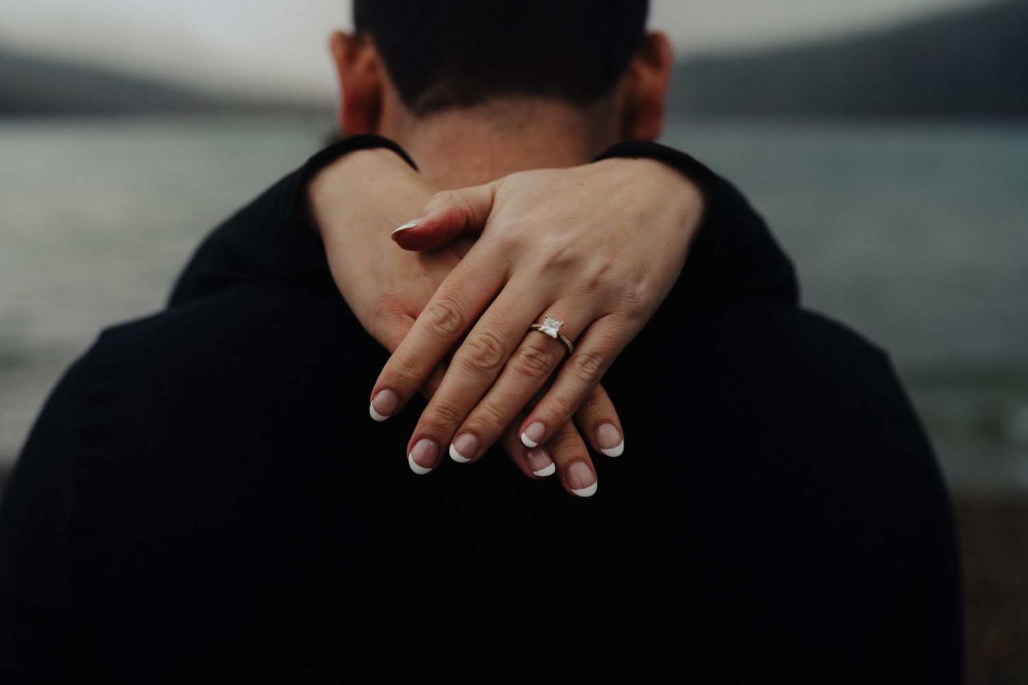 Close up of hands with engagement ring embracing against a lake backdrop.