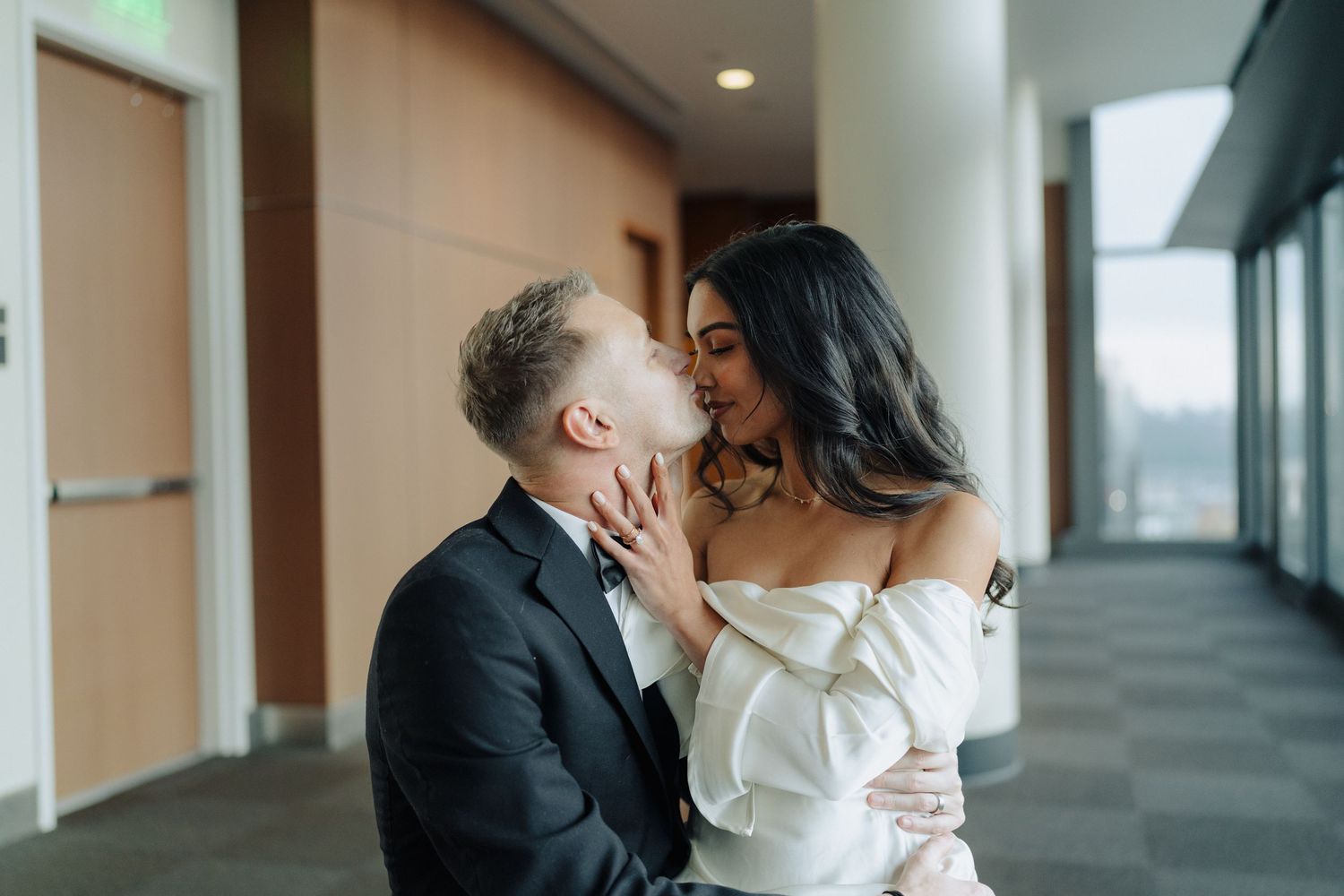 A tender moment between newlyweds in a modern hallway with large windows.