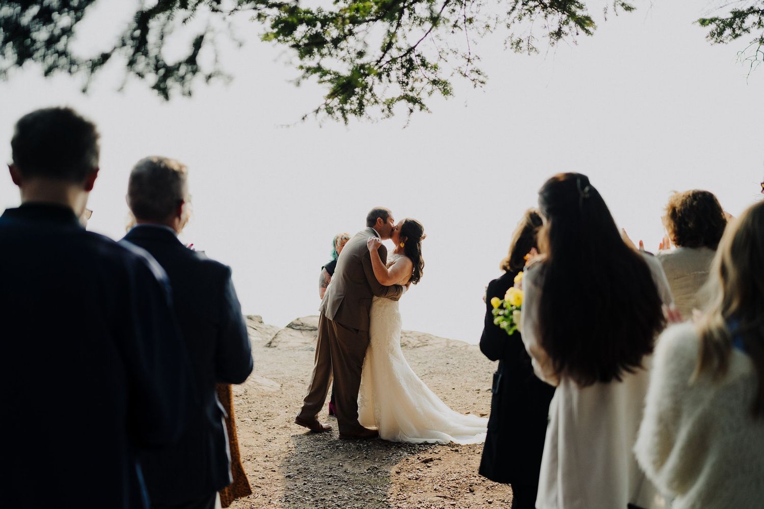 A couple embraces during an intimate outdoor wedding ceremony on a foggy cliffside while guests look on.