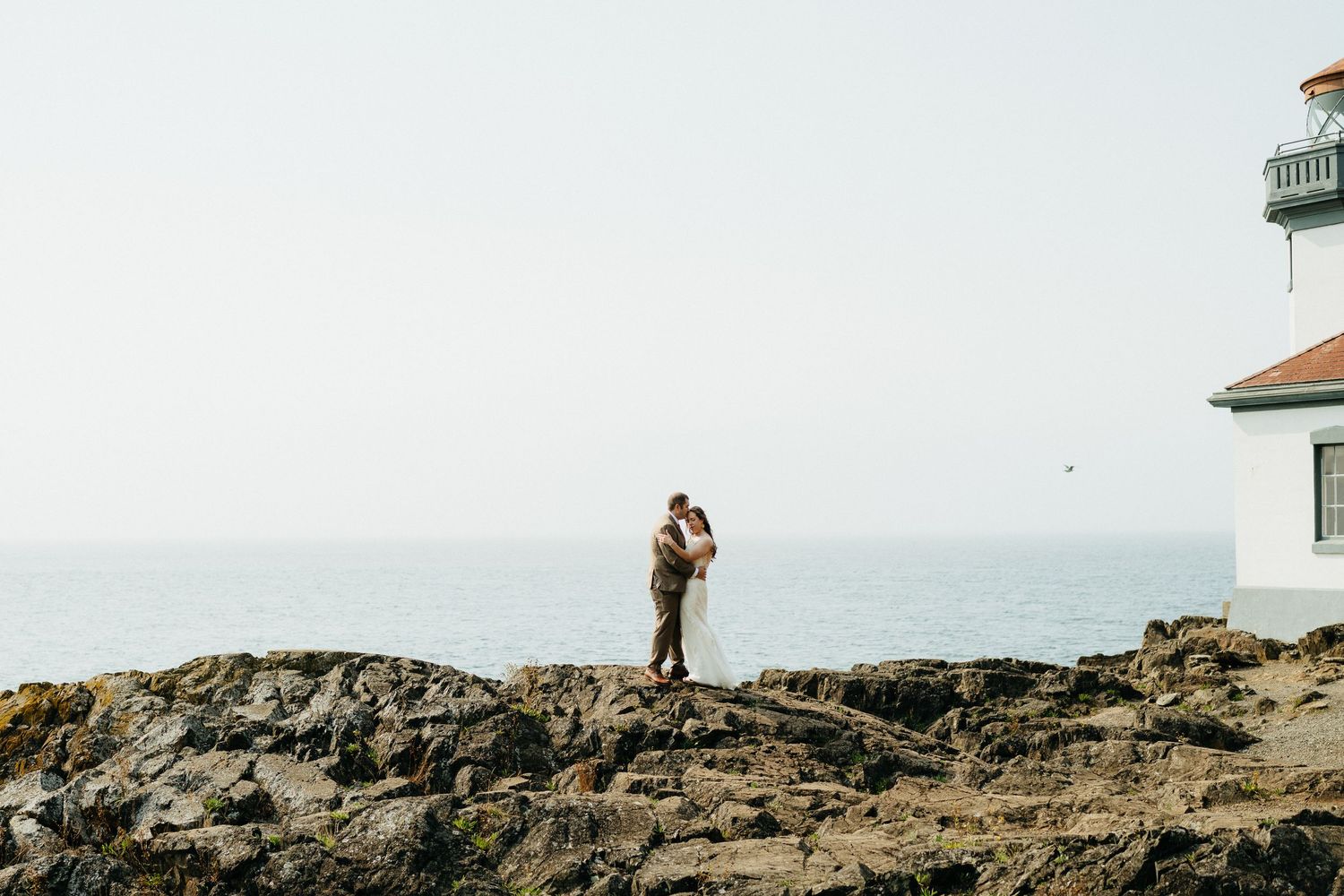 A couple stands on rocky cliffs near a lighthouse overlooking the ocean.