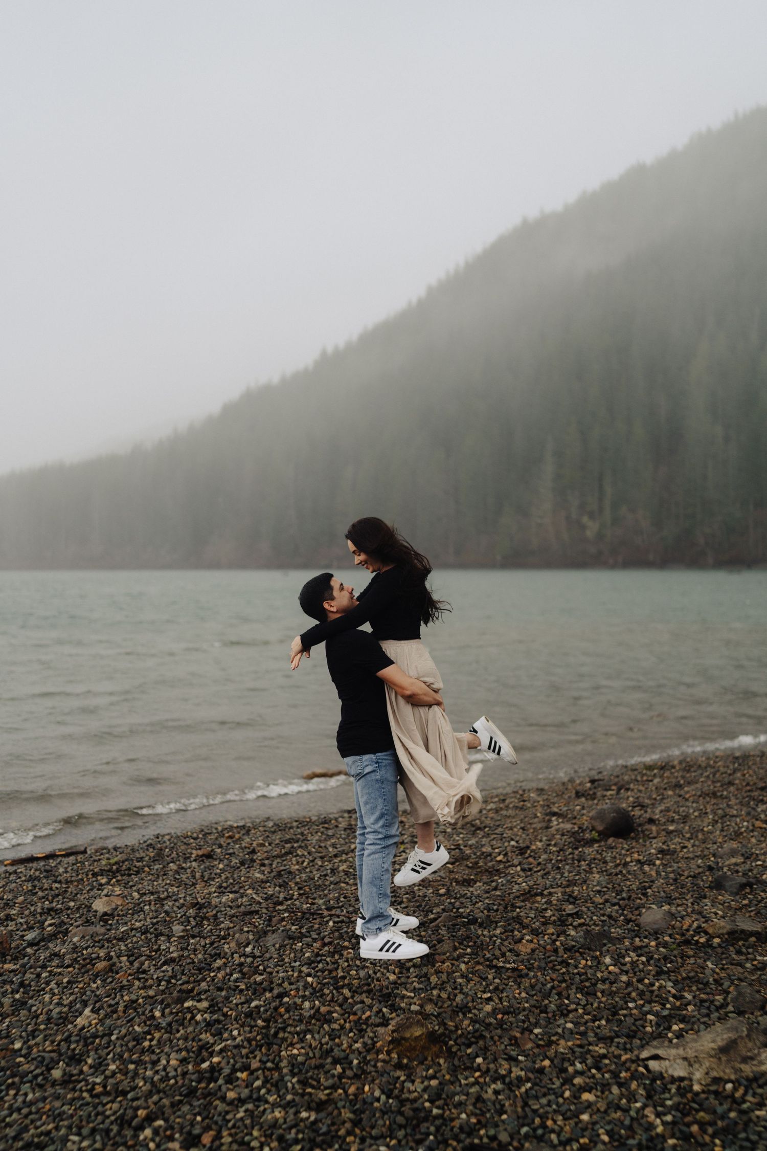 Dancing couple on rocky beach shore with misty mountain landscape and gray skies.