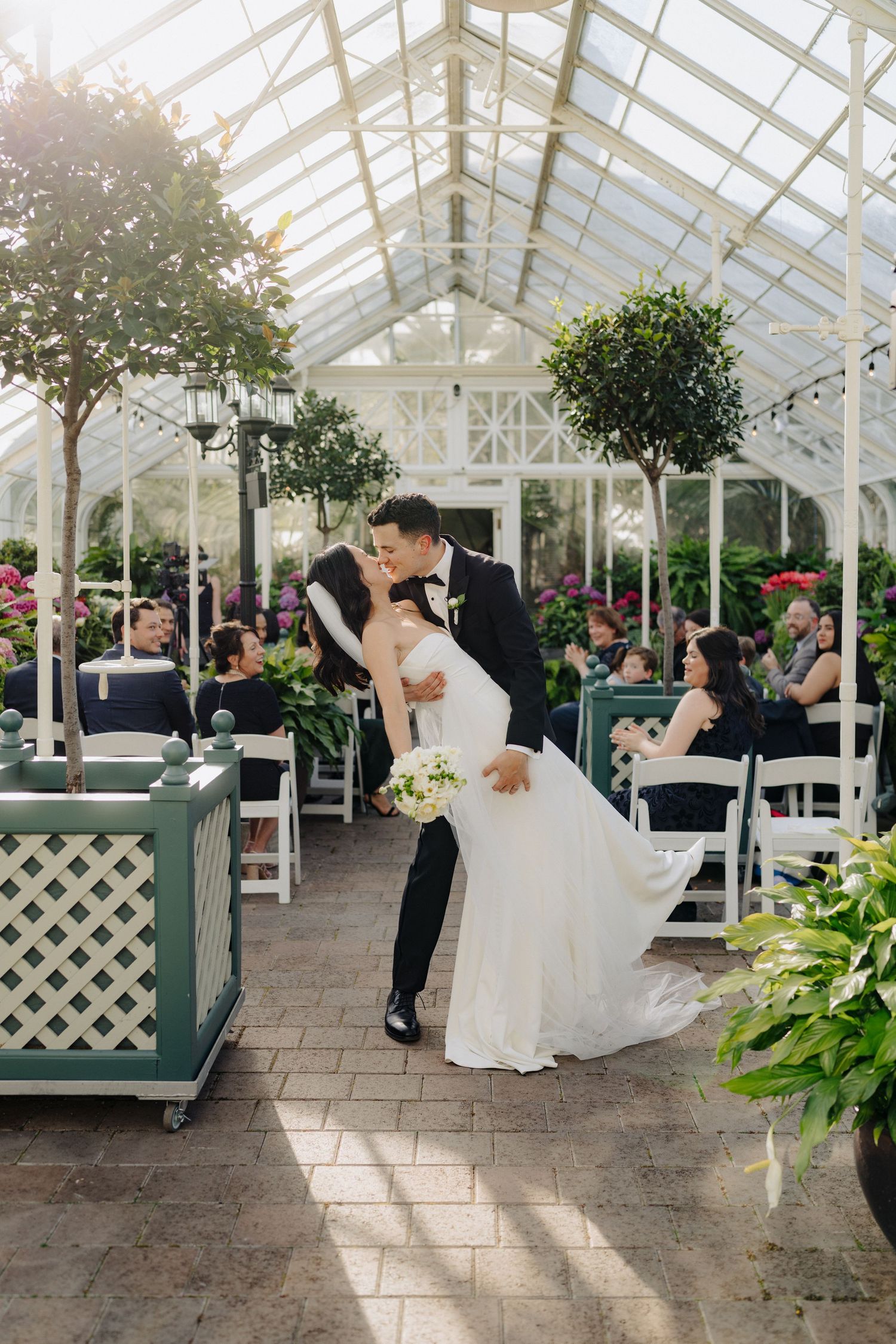 A romantic wedding ceremony in a sunlit greenhouse with white lattice and potted plants creating an intimate atmosphere.