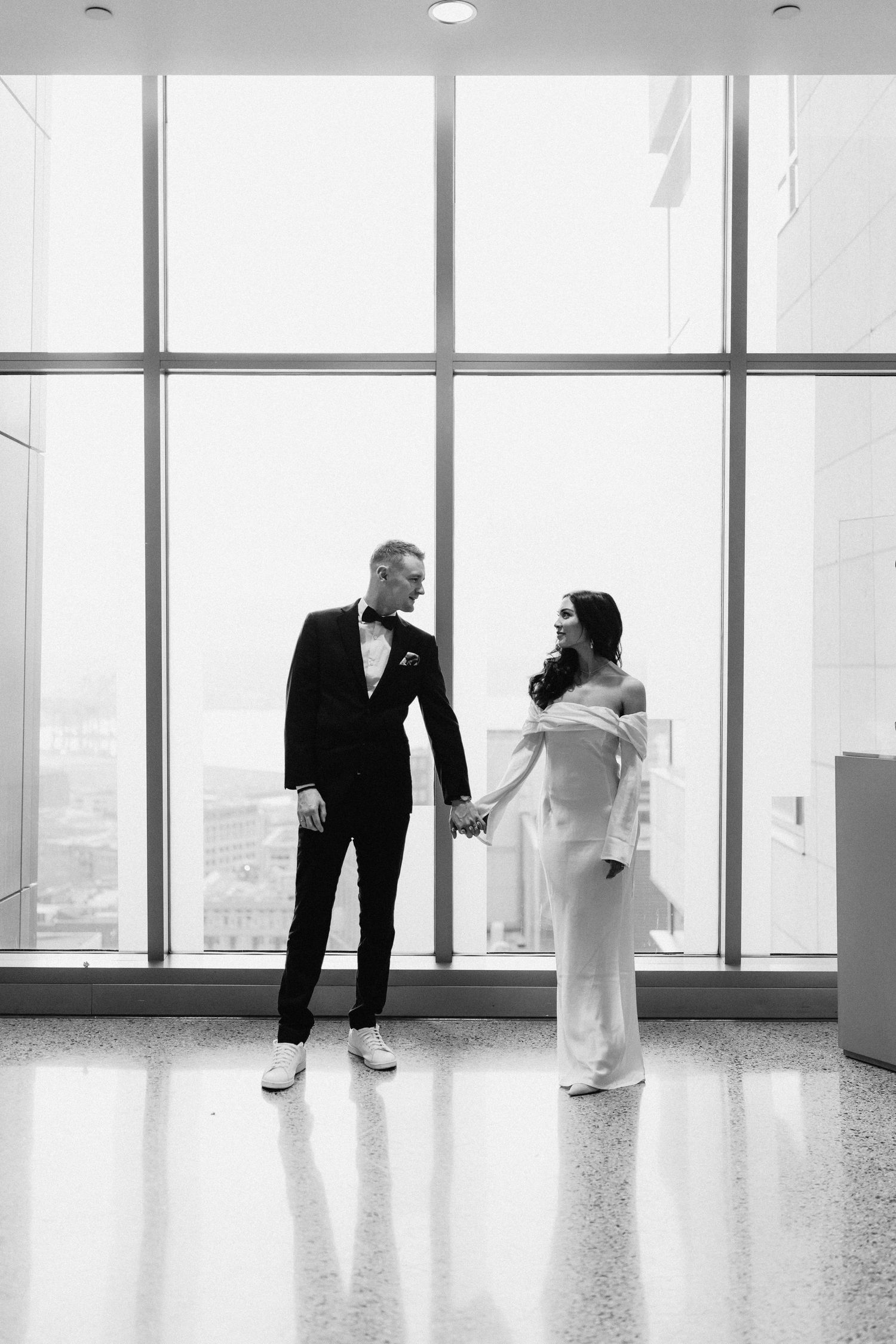 A couple in wedding attire stands together by large floor-to-ceiling windows with a city view.