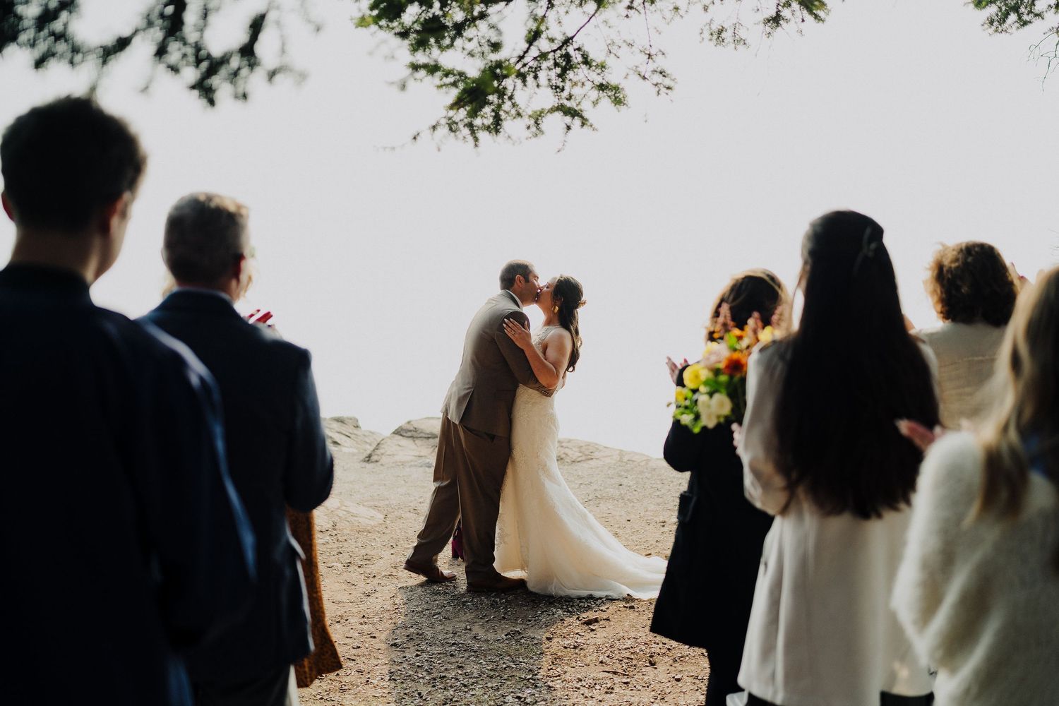 A romantic cliff-side wedding ceremony with onlooking guests silhouetted against a bright misty background.