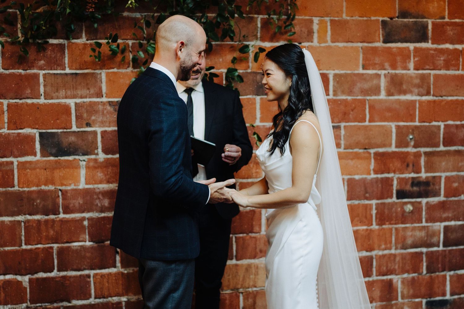 A couple exchanges vows during their wedding ceremony against a rustic red brick wall.