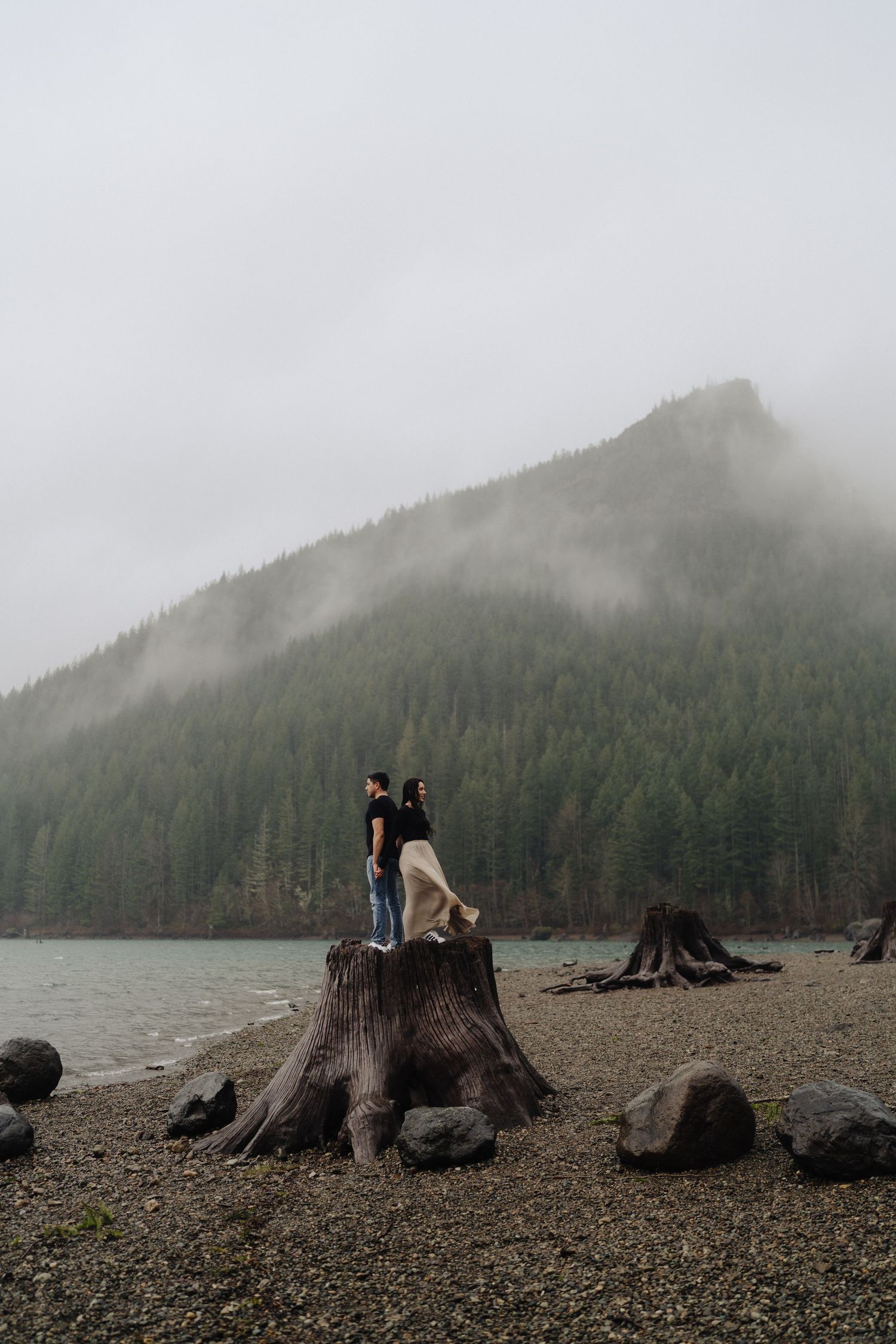 A couple stands on a large tree stump by a misty lake surrounded by evergreen mountains.