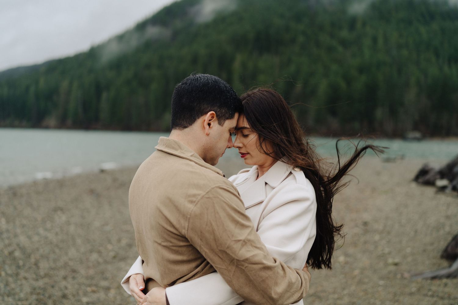 A couple shares a romantic moment on a rocky beach with misty mountains and lake in the background.
