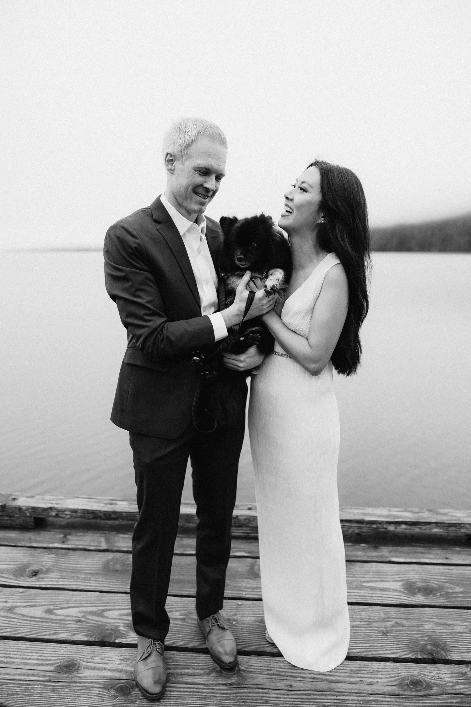 A couple in wedding attire share a joyful moment with their small black dog on a wooden dock by a serene lake.