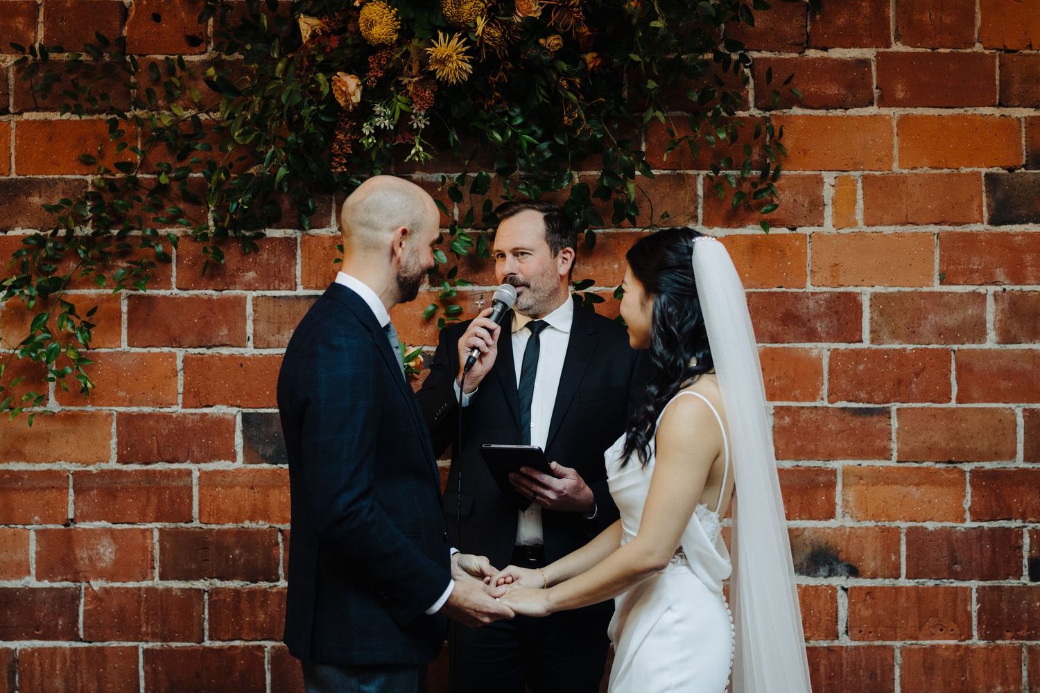A wedding ceremony takes place against a rustic brick wall adorned with climbing vines.