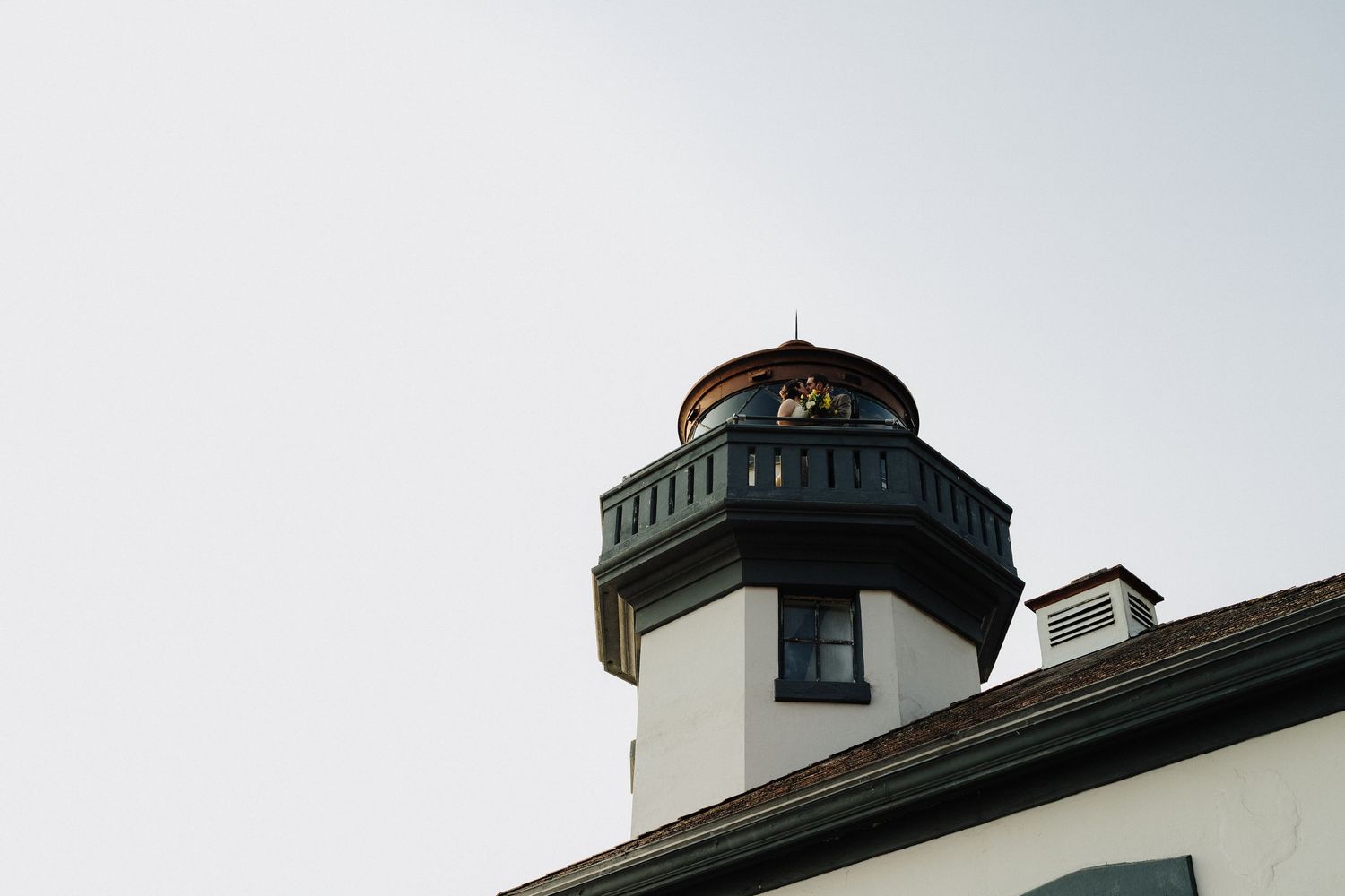 Top portion of a classic white lighthouse with green trim against a pale blue sky.