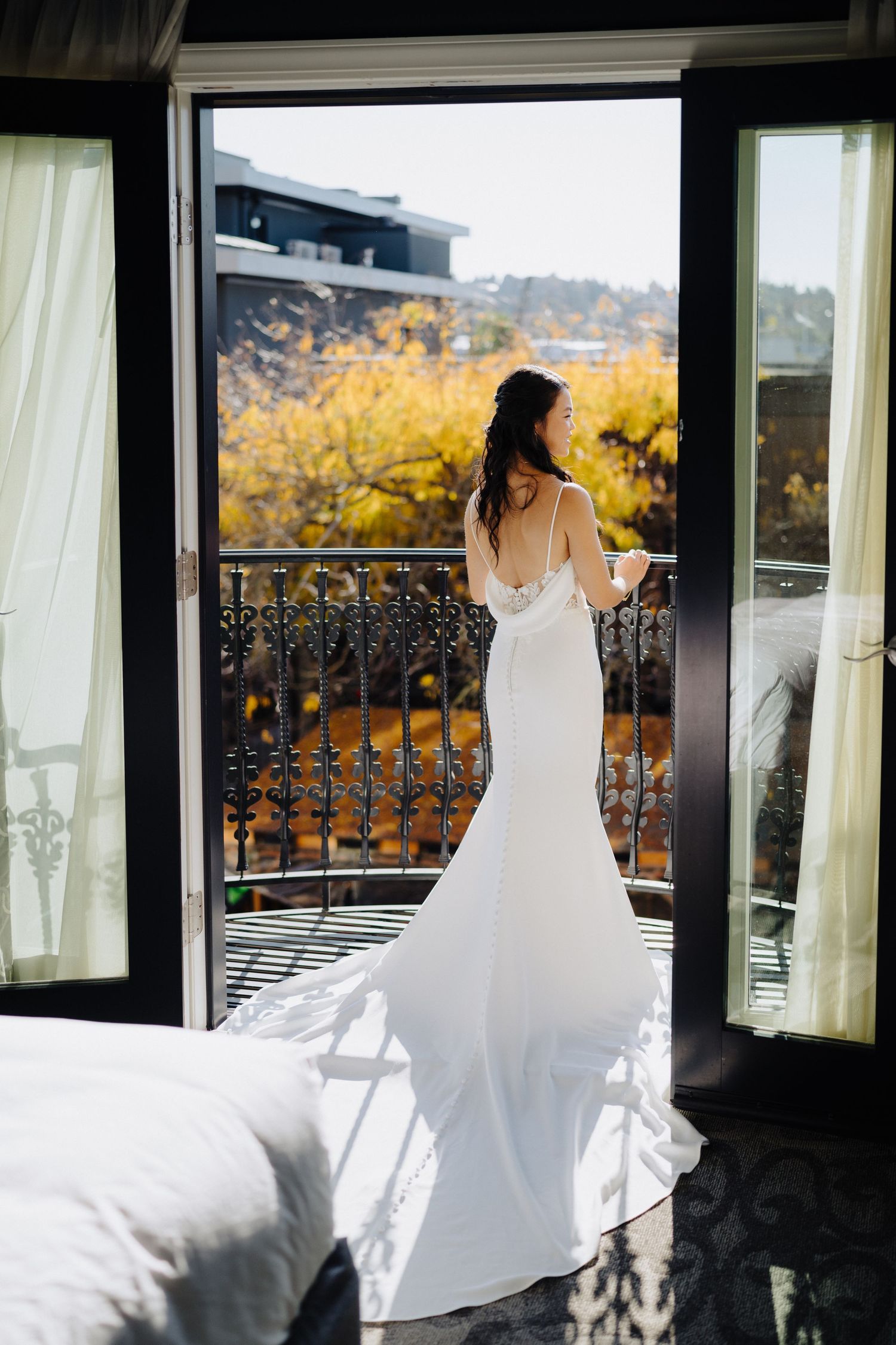 Elegant bride in fitted white dress gazes from balcony overlooking autumn scenery with golden foliage.