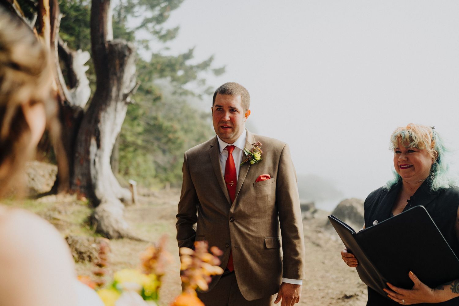 A person in a brown suit with a red tie and boutonniere walks along an outdoor path on a misty day.
