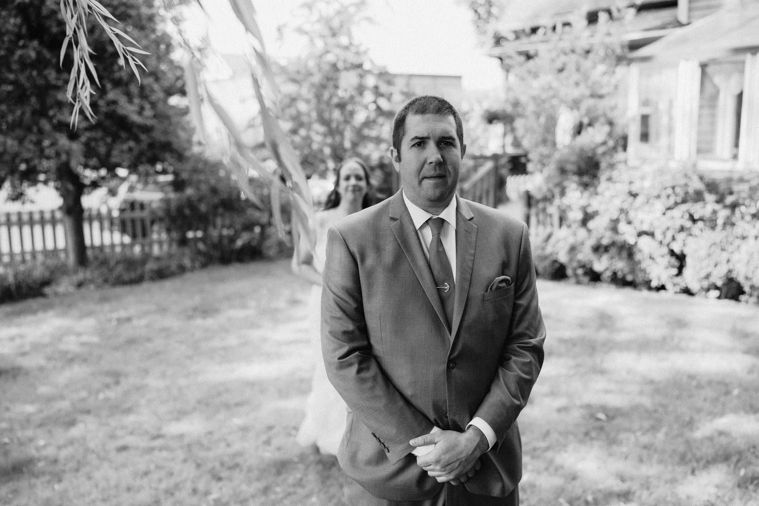 A person stands alone in a formal suit in a garden setting on a sunny day in black and white.