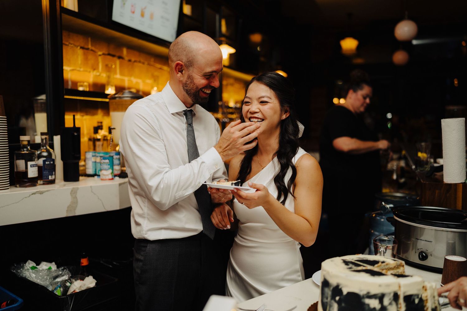 A couple sharing cake at their wedding reception near a bar area.
