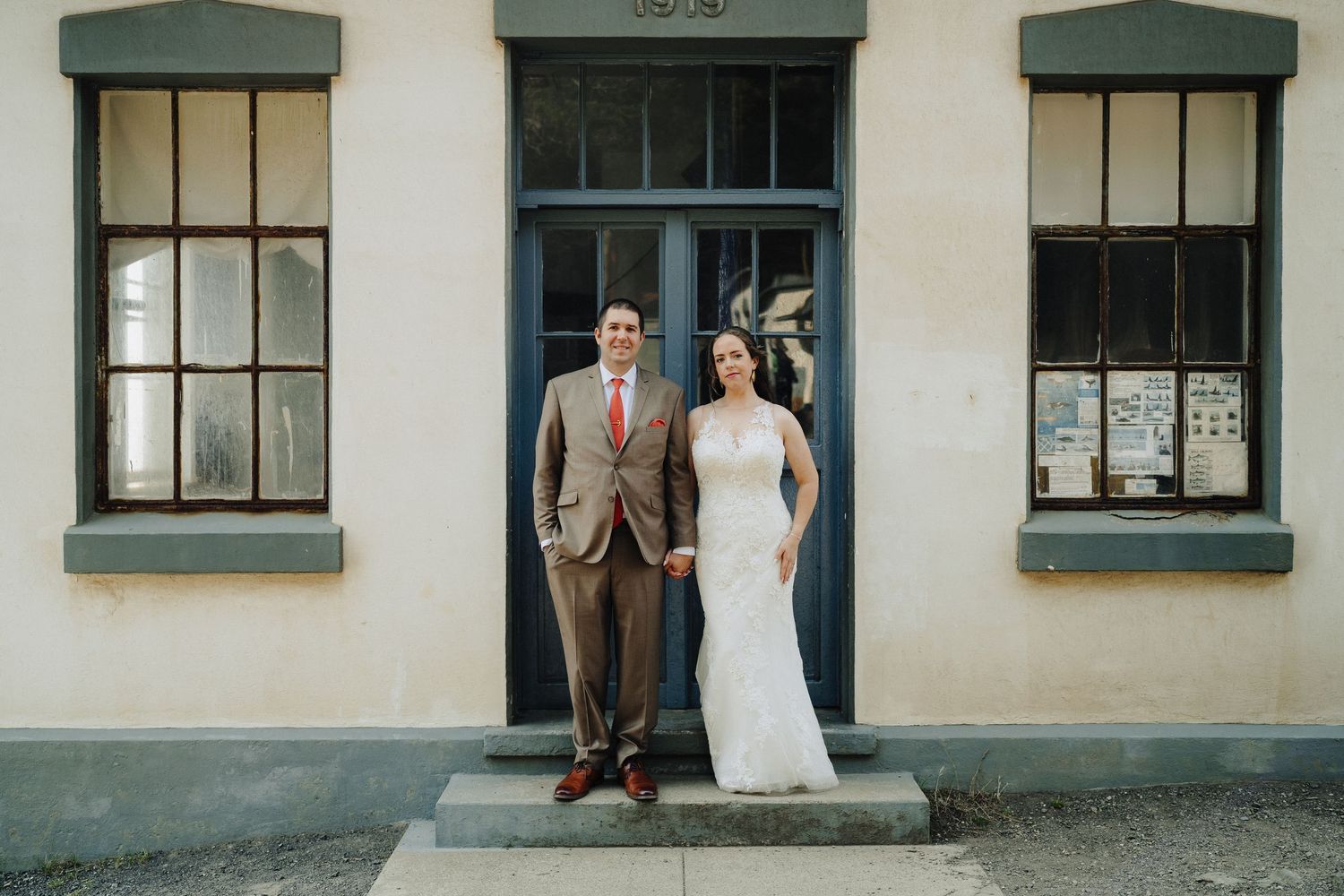 A couple stands in formal wedding attire against a historic building facade with a dark door and green-trimmed windows.