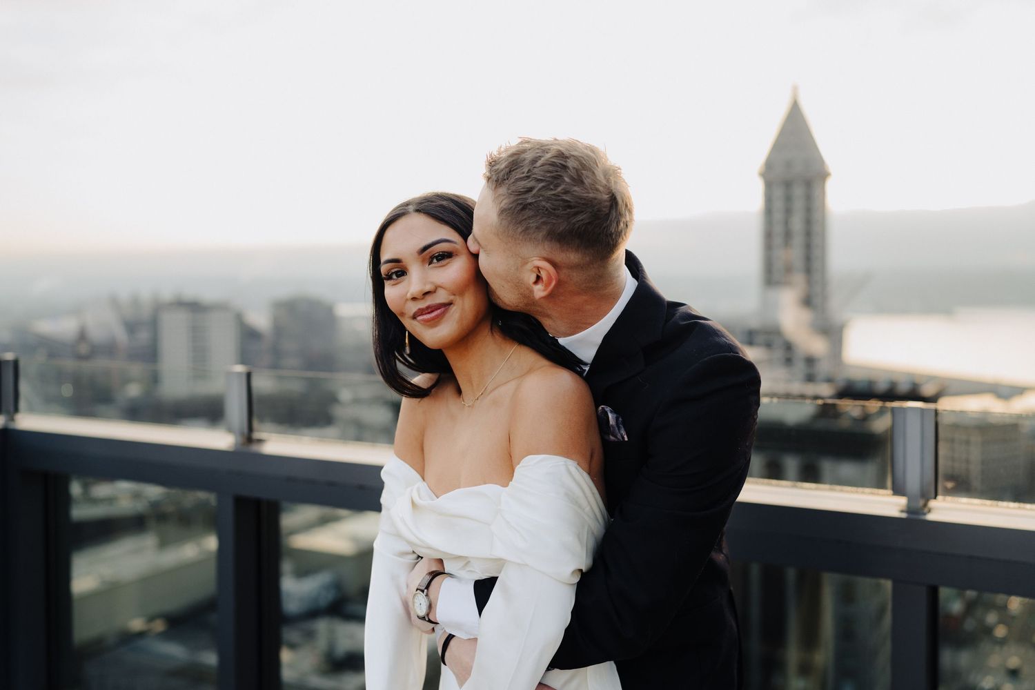A romantic couple embraces on a rooftop terrace with a city skyline and church tower in the background.