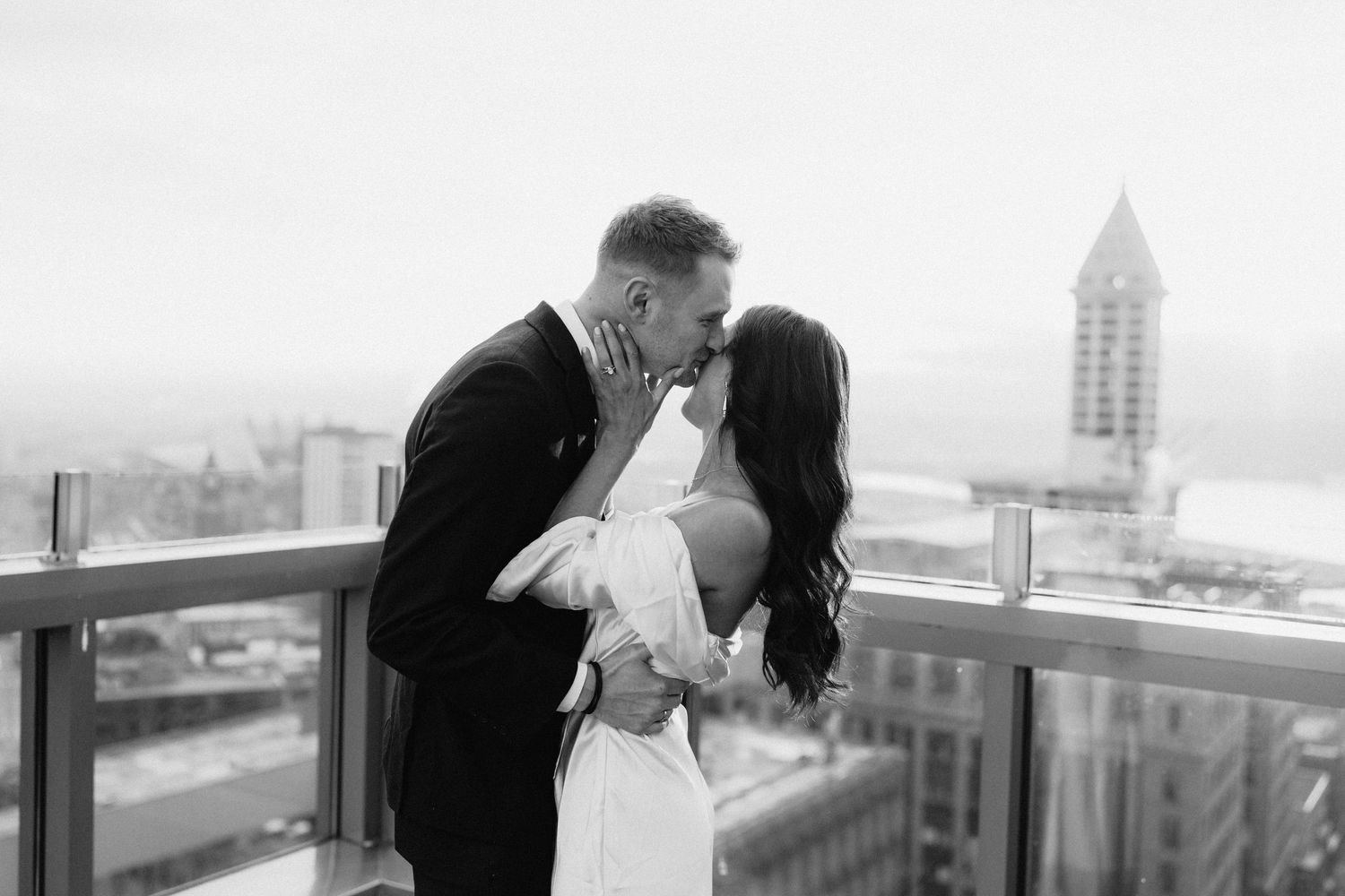 A couple shares a romantic kiss on a rooftop balcony overlooking a cityscape with a prominent tower in the background.