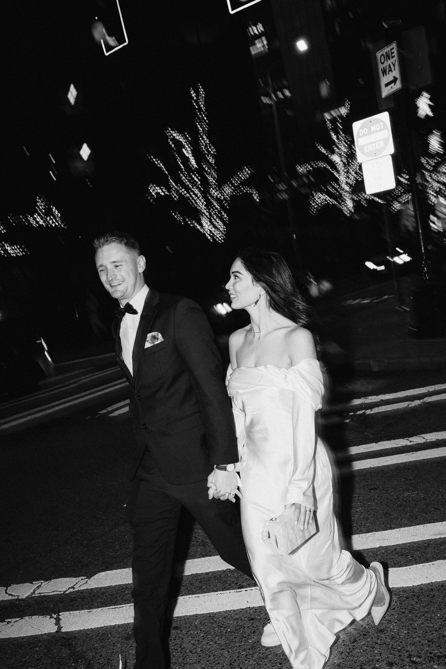 Black and white nighttime street photo of newlyweds walking together in formal attire.