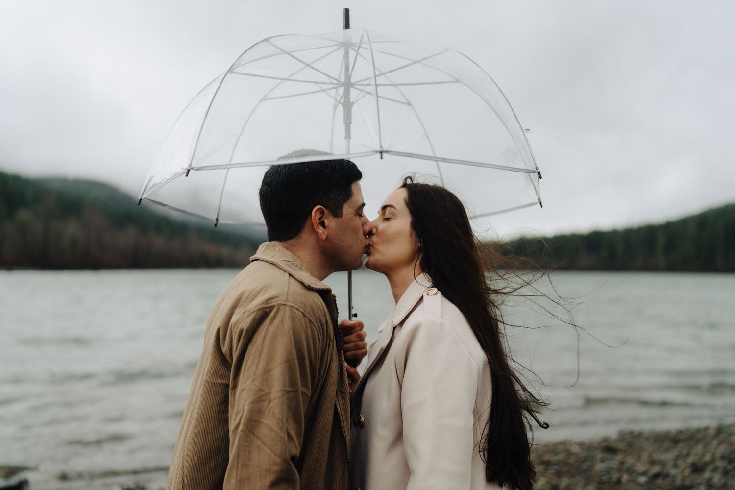 A couple sharing a romantic kiss under a clear umbrella by a misty lake on an overcast day.