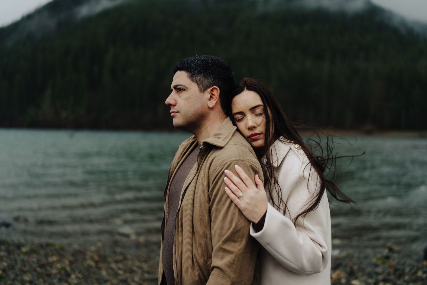 A couple embraces by a misty mountain lake on an overcast day.
