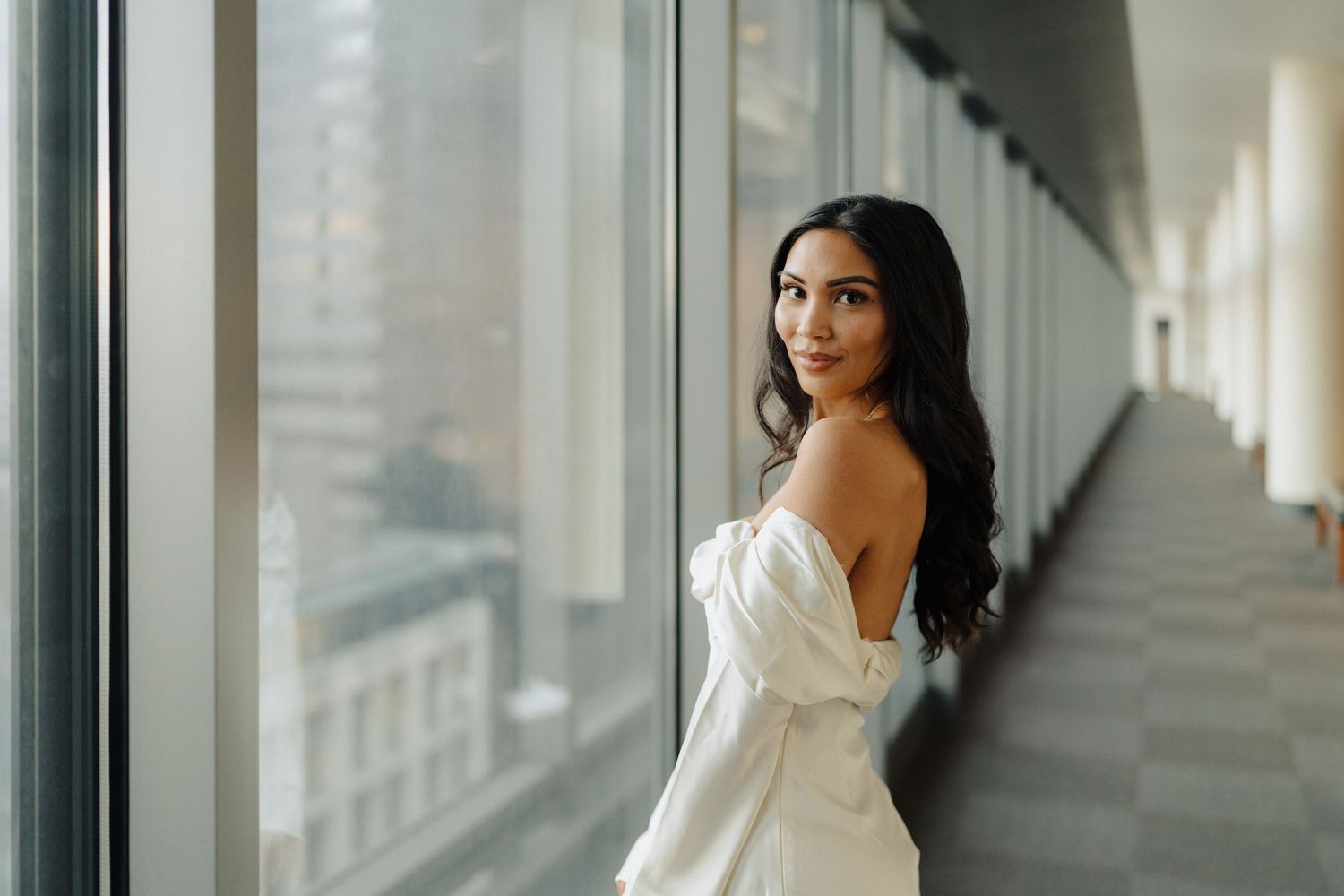 An elegant bride in a white off-shoulder gown poses by a window in a modern building.