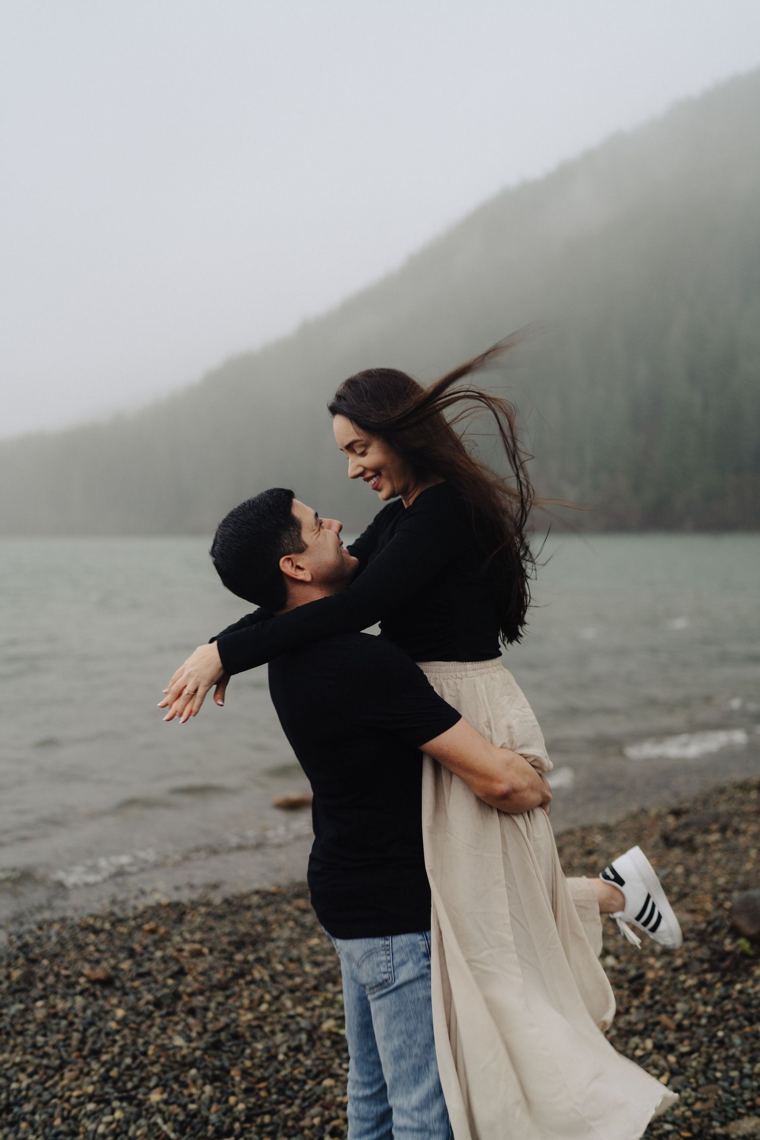 Romantic couple embracing and laughing on pebble beach with foggy mountain scenery.