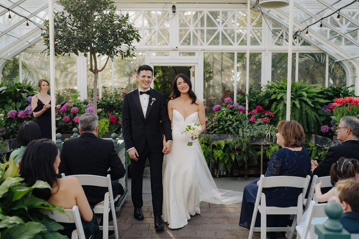 Wedding ceremony setup inside a sunlit greenhouse with white chairs and floral decorations.