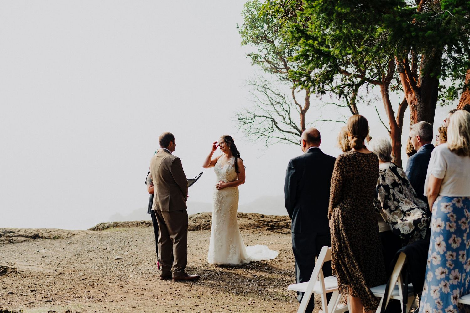 An outdoor wedding ceremony takes place on a scenic cliff overlooking a misty landscape with guests gathered under trees.