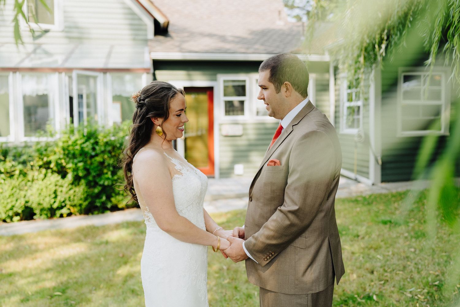 A couple shares a tender moment holding hands outdoors in front of a house with lush greenery.