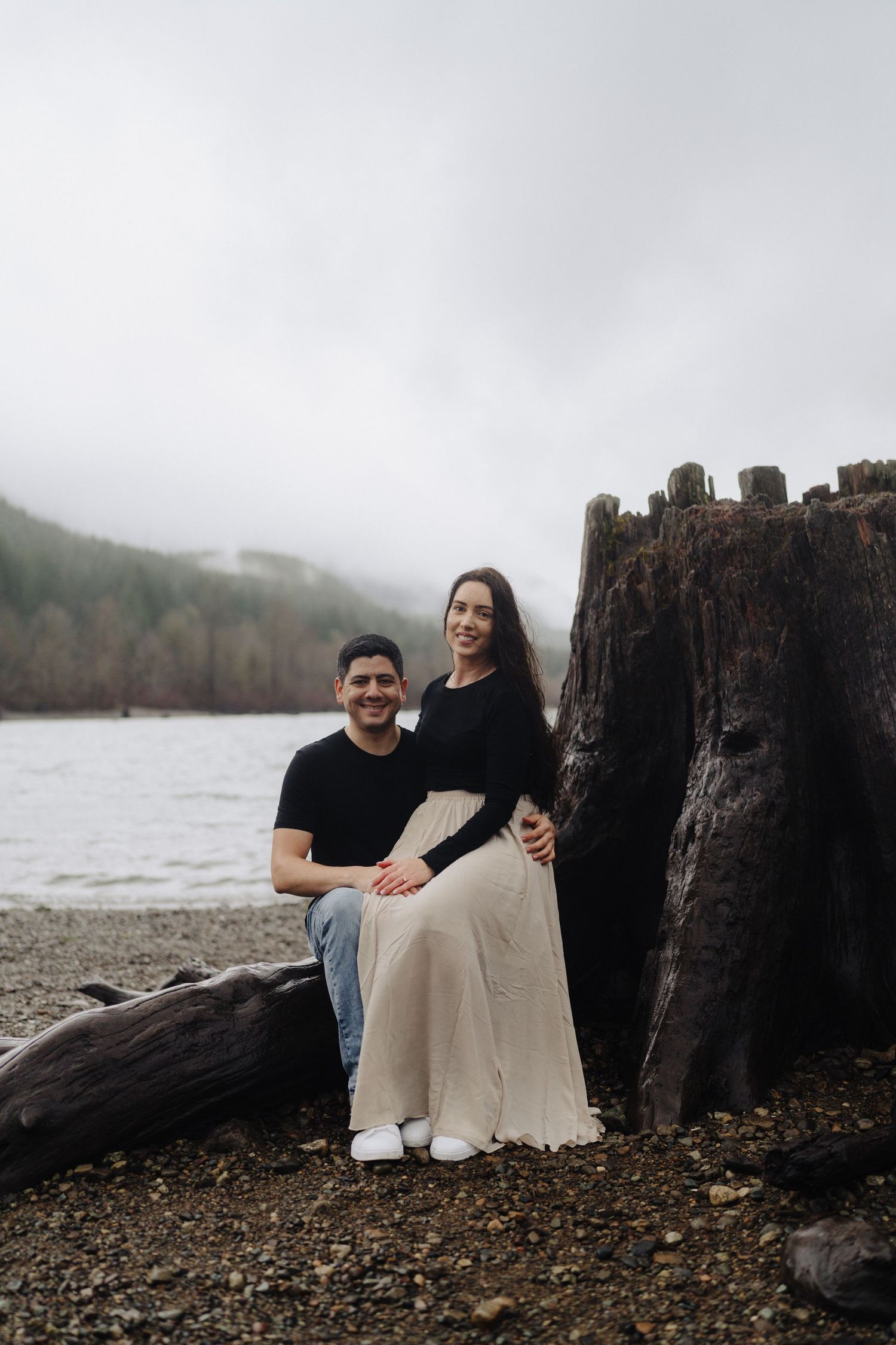 Couple sitting together on rocky beach shore with misty mountain backdrop and dark waters.