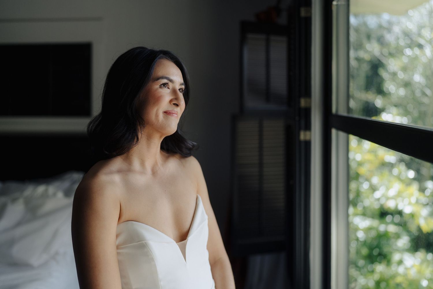 Woman in a white wedding dress gazes thoughtfully out a bright window.