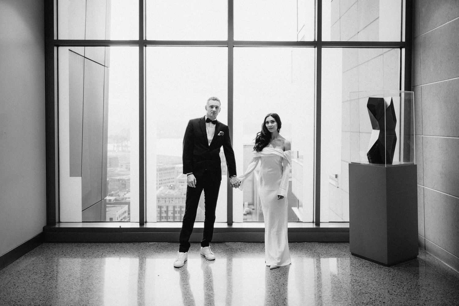 A black and white photo of a couple standing in a modern gallery space with large windows.